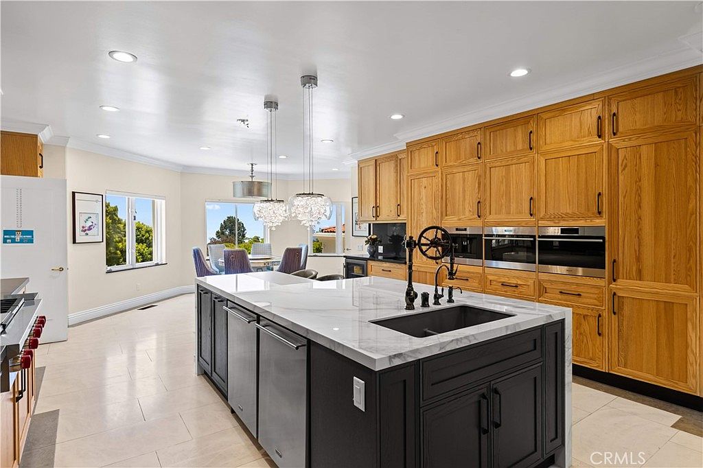 This spacious kitchen features a large central island with a dark base and a light stone countertop, complemented by extensive light wood cabinetry along the wall. The room is illuminated by recessed lighting and two decorative pendant lights hanging over the island, while a dining area is visible in the background through large windows. The neutral tile flooring and clean lines create a bright, open, and functional culinary space.