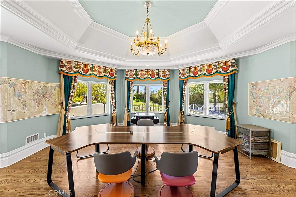This bright and spacious office features a large, custom-shaped wooden desk positioned centrally under a decorative tray ceiling with a classic chandelier. The room is painted in a soft blue hue and includes two large wall maps, providing an educational or professional atmosphere. Large windows with patterned valances and drapes allow for ample natural light, creating an inviting and functional workspace.