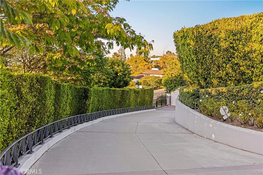 This image captures a paved driveway leading up to a residential property, framed by lush, manicured hedges and mature trees. The perspective is from the base of the incline, looking upward toward the home, emphasizing the private and secluded nature of the entrance. A decorative metal railing runs along the left side, adding a structured element to the verdant landscape.