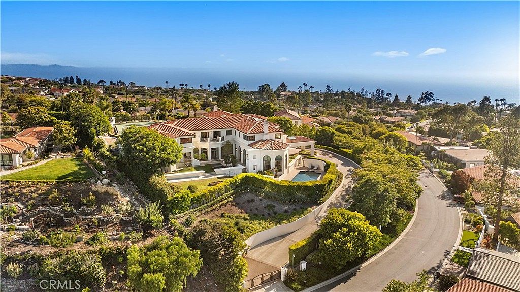 This high-angle aerial view captures a sprawling Mediterranean-style estate nestled in a coastal landscape. The property features a multi-level residence with a terracotta-tiled roof, a private swimming pool, and lush, manicured landscaping. The expansive grounds overlook the ocean in the distance, emphasizing the home's prime location and luxurious scale.