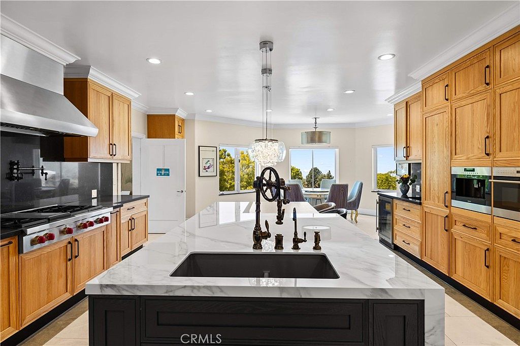 This spacious, open-concept kitchen features a large central island with a dark base and a light-toned stone countertop, serving as the focal point of the room. The perimeter is lined with light wood cabinetry, stainless steel appliances, and a dark stone backsplash, creating a high-contrast aesthetic. A bright dining area is visible in the background, illuminated by natural light from large windows that offer a view of the outdoors.
