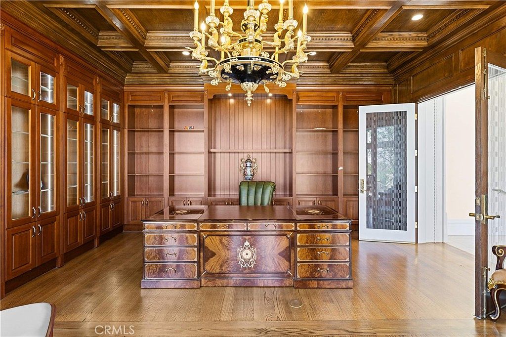 This sophisticated home office features extensive custom wood cabinetry and built-in shelving that spans the back wall. A large, ornate wooden desk sits centrally in the room beneath a grand, multi-tiered crystal chandelier hanging from a coffered ceiling. The space is finished with warm-toned wood flooring and a classic green leather desk chair, creating an atmosphere of traditional elegance and professional refinement.