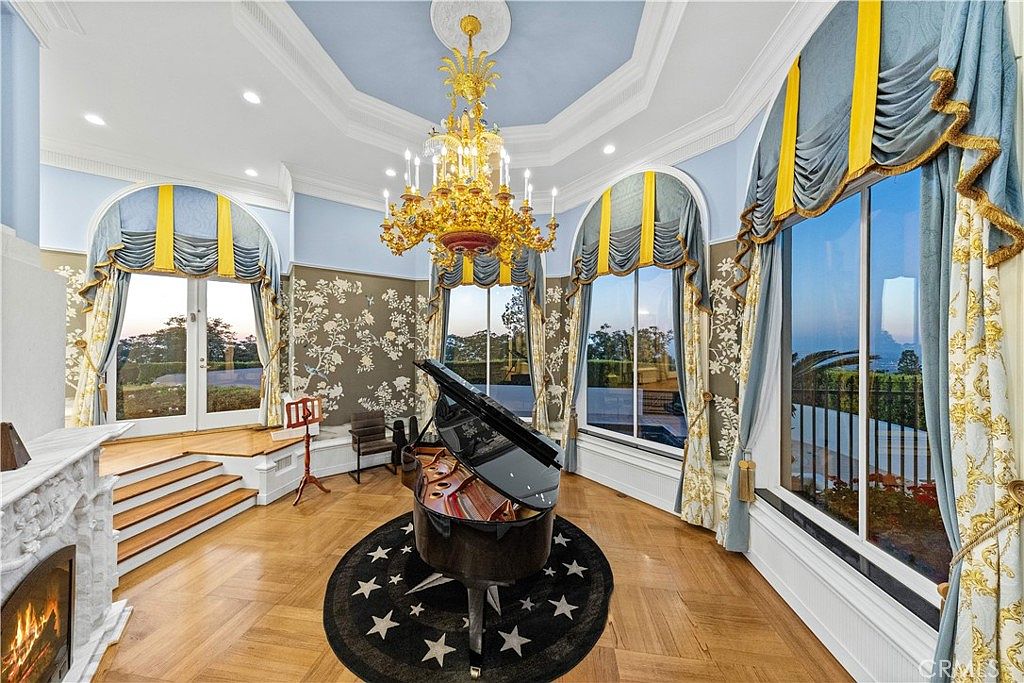 This elegant formal living space features a grand piano centered on a star-patterned rug beneath an ornate gold chandelier. The room is defined by tall arched windows with blue and yellow drapery, light wood flooring, and walls adorned with intricate floral wallpaper. A fireplace with a detailed white surround adds a touch of classic sophistication to the bright, airy atmosphere.