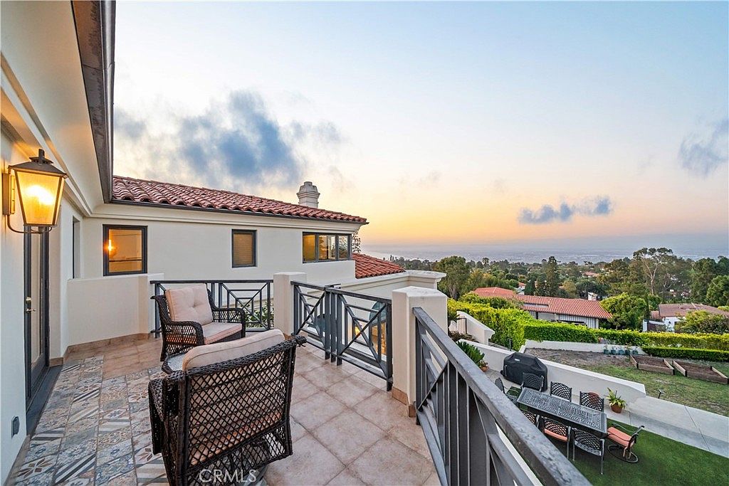 This elevated balcony offers a scenic view of the surrounding landscape and distant ocean horizon under a soft sunset sky. The space is furnished with comfortable wicker-style seating and features decorative patterned tile flooring, enclosed by a dark metal railing. Below the balcony, a well-maintained backyard with a dining area and lawn provides a secondary outdoor living space.