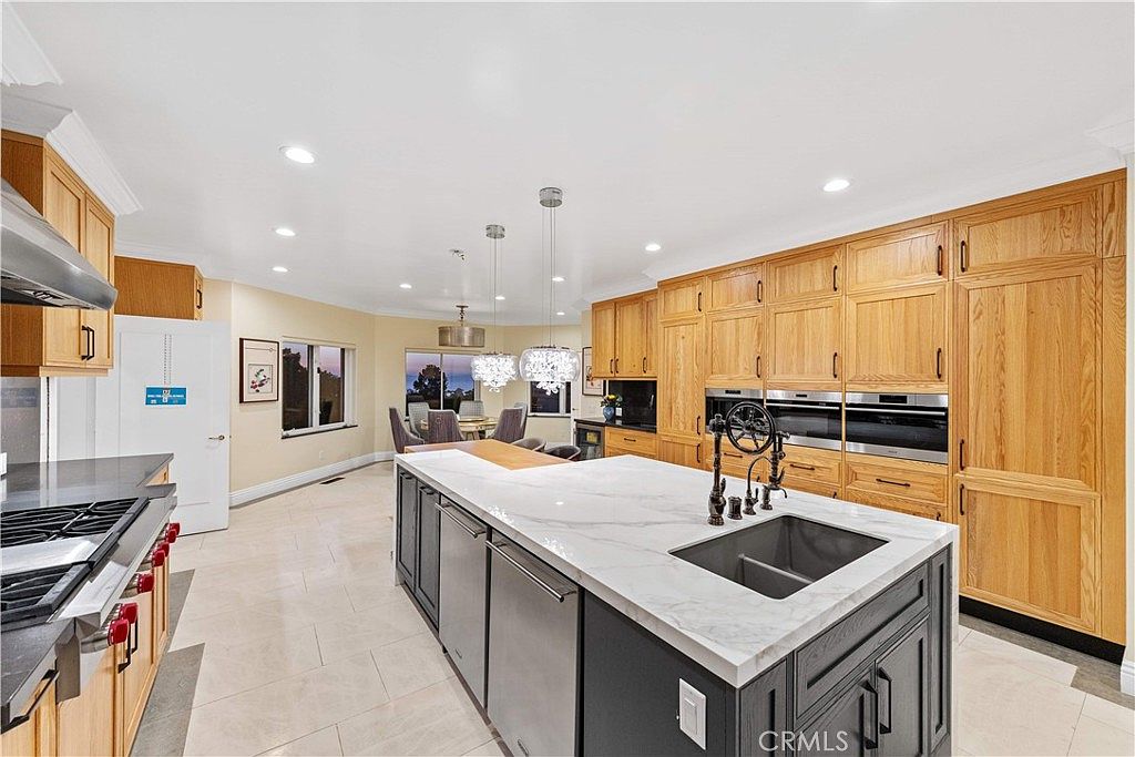 This spacious kitchen features a large central island with a dark base and a light-toned stone countertop, complemented by extensive light wood cabinetry throughout. The room is equipped with built-in stainless steel appliances and is illuminated by recessed lighting and decorative pendant fixtures. A dining area is visible in the background, creating an open-concept flow that looks out toward a windowed view.