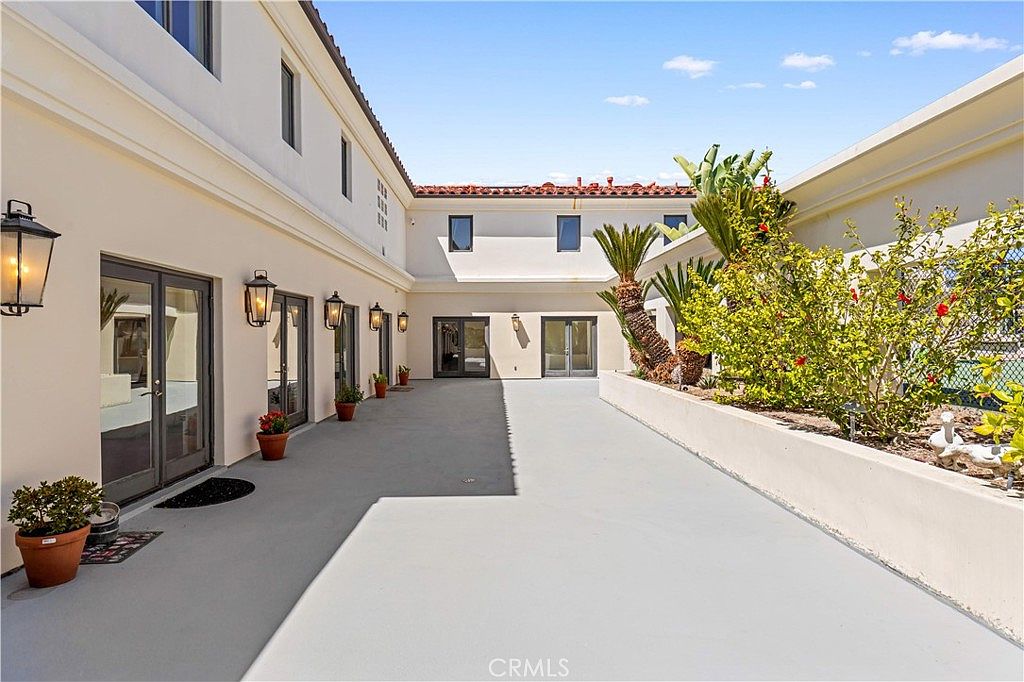 This image captures a spacious, paved courtyard area featuring a Mediterranean-inspired architectural style with smooth, light-colored walls and a red-tiled roofline. A series of glass doors with dark frames line the left side of the courtyard, accented by wall-mounted lanterns and potted plants. To the right, a raised planter bed filled with lush greenery and palm trees adds a natural element to the clean, minimalist outdoor space.