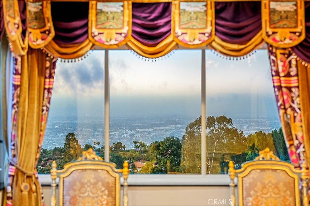 This interior view features a formal dining area framed by ornate, heavy purple and gold valance curtains that add a touch of traditional elegance. Large windows provide a panoramic, elevated view of the sprawling city landscape and surrounding greenery below. In the foreground, the tops of two upholstered chairs with decorative wood trim are visible, suggesting a sophisticated and classic interior design style.