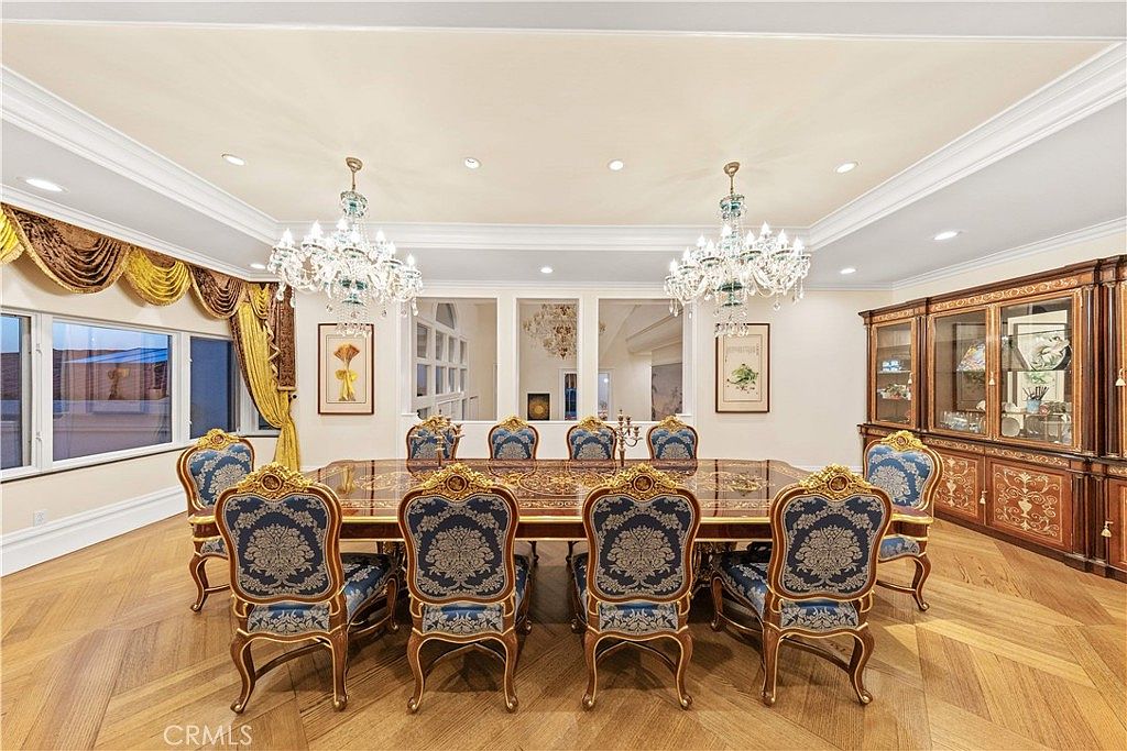 This formal dining room features a large, ornate rectangular table surrounded by ten high-backed chairs upholstered in blue patterned fabric. The space is illuminated by two grand crystal chandeliers hanging from a tray ceiling, complemented by a large wooden china cabinet against the back wall. The room is finished with light wood flooring arranged in a herringbone pattern and elegant gold-toned window treatments.