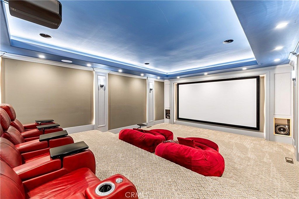 This dedicated home theater room features a large projection screen mounted on a wall with integrated speakers and recessed lighting. The space is furnished with multiple rows of plush red leather reclining chairs and large, comfortable red floor cushions for casual seating. The room is finished with a light-colored carpet and a blue-painted ceiling, creating a cozy and immersive cinematic environment.