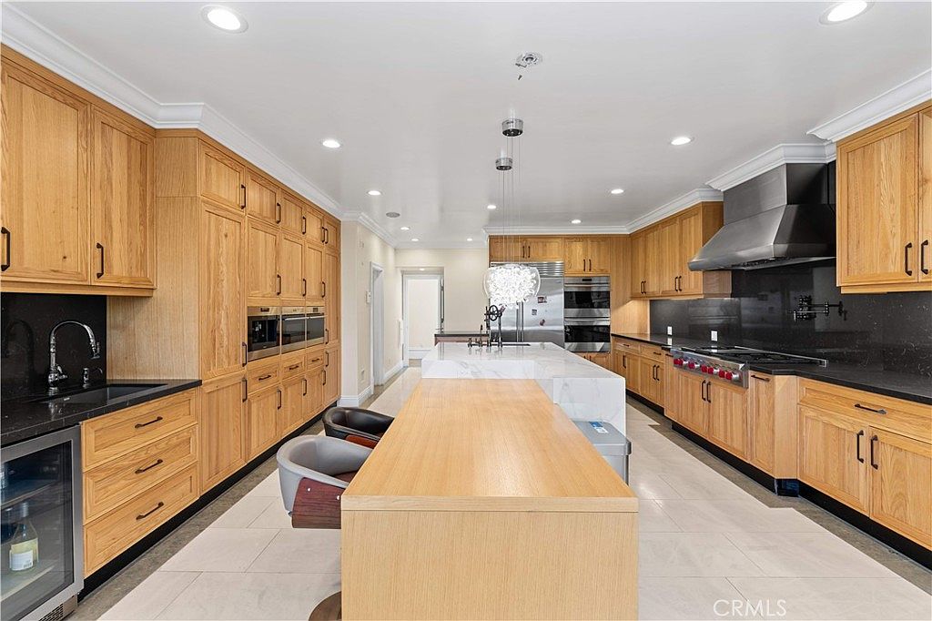 This spacious, modern kitchen features extensive light wood cabinetry and a large central island with a wood-topped breakfast bar. The space is equipped with high-end stainless steel appliances, including a professional-style range and built-in wall ovens, complemented by dark stone countertops and a matching backsplash. Recessed lighting and a decorative pendant fixture illuminate the room, which is finished with light-colored tile flooring.