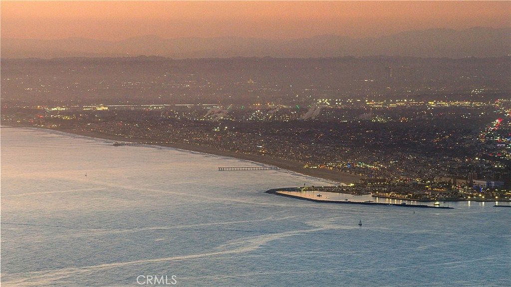 This high-altitude aerial view captures a sprawling coastal landscape at dusk, featuring a long stretch of sandy beach meeting the calm ocean waters. A prominent pier extends into the sea, while the shoreline is lined with a dense urban development illuminated by city lights. The perspective provides a cinematic, wide-angle look at the property's proximity to the coastline and the surrounding community.
