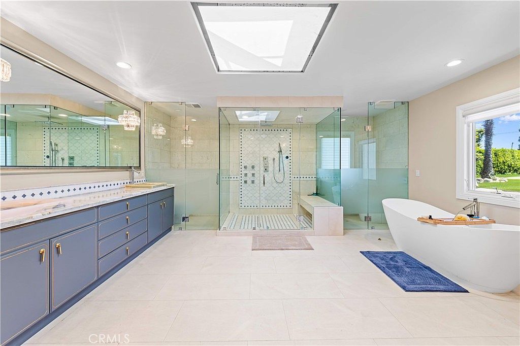 This spacious primary bathroom features a large, central glass-enclosed shower with a built-in bench and intricate tile detailing. To the left, a long vanity with blue cabinetry and a wide mirror provides ample storage, while a freestanding soaking tub sits to the right near a window. The room is bright and airy, enhanced by a large skylight that floods the space with natural light.