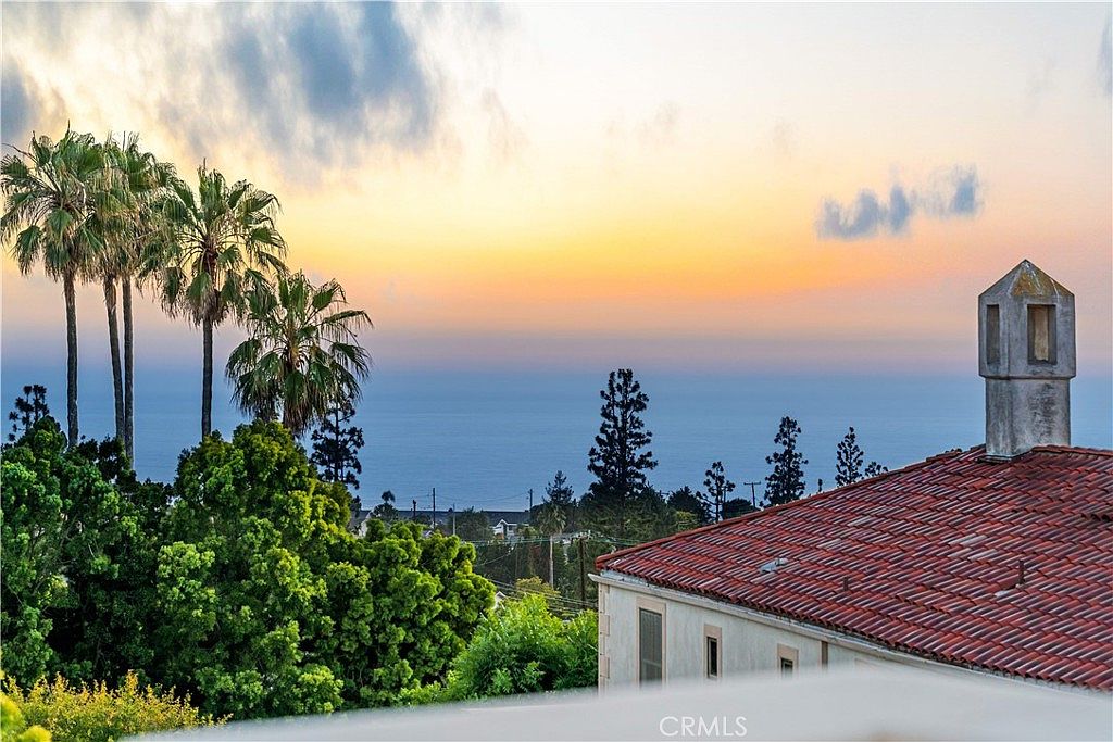 This elevated perspective captures a stunning sunset view over the ocean from a residential property. The foreground features a portion of a tiled roof with a prominent chimney, while lush green trees frame the horizon. The scene evokes a serene and luxurious atmosphere, highlighting the home's scenic coastal location.