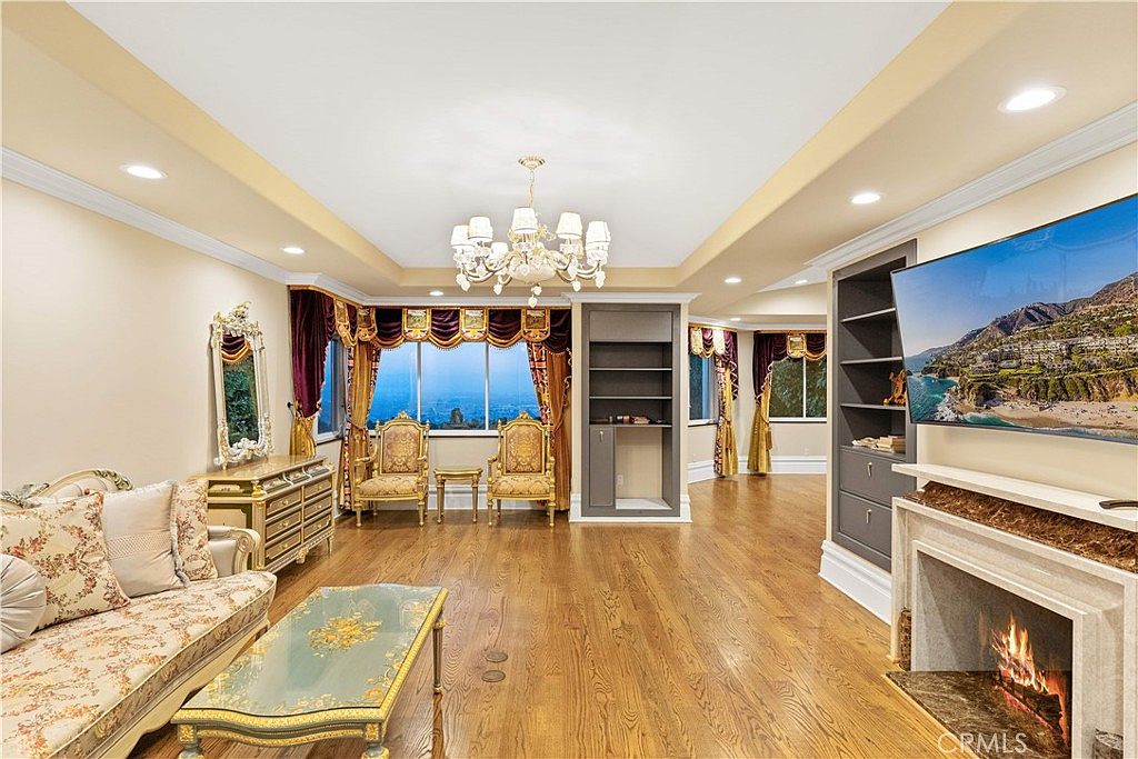 This elegant living room features polished light wood flooring and a sophisticated, traditional aesthetic. The space is anchored by a fireplace with a stone surround and a large television mounted above, while a crystal chandelier hangs from the tray ceiling. Large windows with ornate valances frame a scenic outdoor view, and the room is furnished with a patterned sofa and matching gold-toned accent chairs.