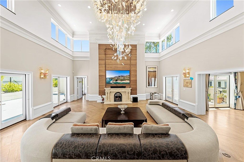 This expansive living room features a dramatic, multi-tiered crystal chandelier suspended from a high ceiling, serving as the focal point of the space. A large, curved sectional sofa faces a central coffee table and a stone-clad chimney breast with a built-in television. The room is framed by large windows and glass doors that provide abundant natural light and views of the surrounding outdoor areas, all set against a backdrop of light-toned walls and herringbone-patterned wood flooring.