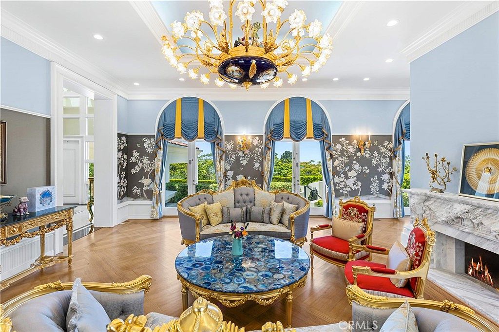 This opulent living room features a grand crystal chandelier, ornate gold-trimmed furniture, and a striking blue agate-topped coffee table. The space is defined by light blue walls, intricate floral wallpaper accents, and large arched windows draped in elegant blue and yellow fabric. A fireplace with a detailed stone mantel adds a focal point to the room, which is finished with polished light wood flooring.
