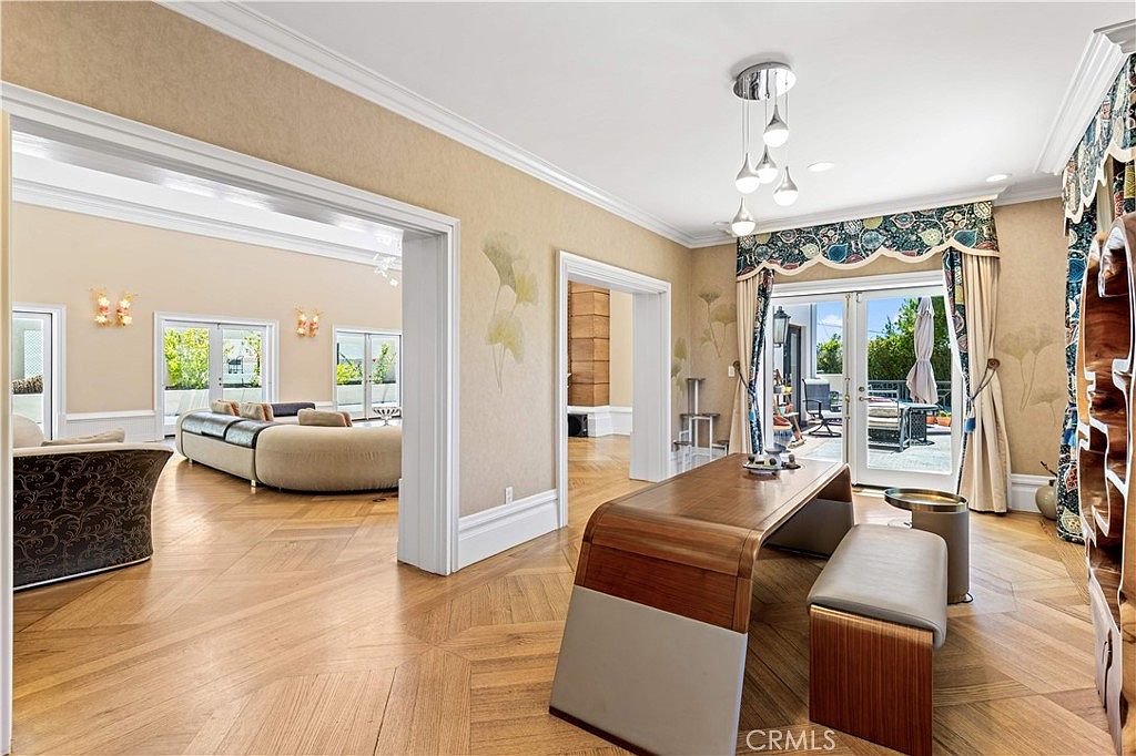 This elegant dining area features a unique, curved wooden table paired with a matching bench, set against a backdrop of light-toned walls with subtle botanical wallpaper patterns. The room showcases beautiful herringbone-patterned wood flooring and opens into a spacious living area through a wide, cased doorway. Large glass doors with decorative valances provide access to an outdoor patio, filling the space with natural light.