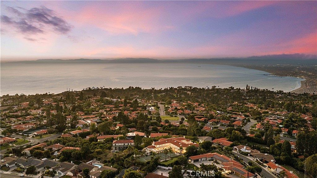This high-angle aerial view captures a sprawling coastal residential neighborhood at sunset, featuring a prominent estate with a tennis court and a large, multi-winged residence. The perspective looks out over the expansive ocean horizon, highlighting the prime location and the lush, tree-lined streets of the community. The warm, soft light of the setting sun casts a serene glow over the entire landscape, emphasizing the property's desirable coastal setting.