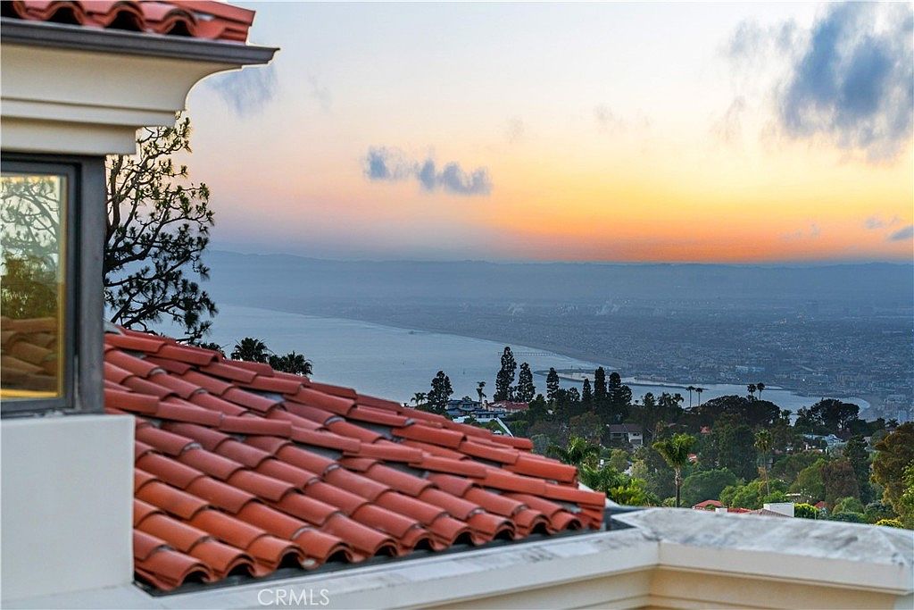 This elevated perspective captures a stunning sunset view over a coastal landscape from a balcony featuring a classic terracotta-tiled roof. The foreground showcases the textured red tiles and a portion of a building's exterior wall, while the background reveals a sweeping panorama of the ocean, coastline, and distant hills under a soft, golden-hued sky. The composition emphasizes the property's prime location and scenic outlook, creating a serene and luxurious atmosphere.