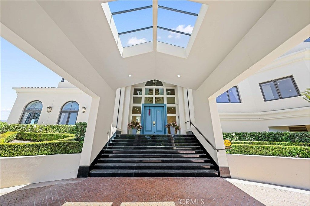 This grand entryway features a covered portico with a glass-paneled roof that allows natural light to flood the space. A set of wide, dark stone steps leads up to a prominent set of double doors painted in a soft blue hue. The surrounding architecture is characterized by clean white walls, arched windows, and manicured hedges, creating a sophisticated and welcoming first impression.