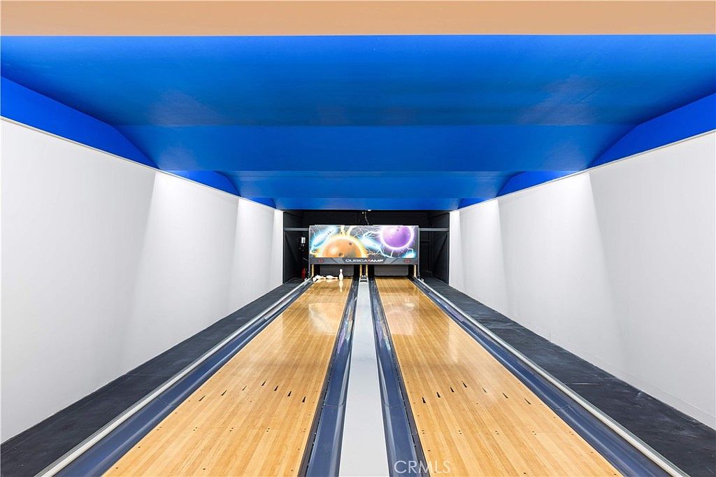 This image showcases a private, two-lane indoor bowling alley featuring polished light wood lanes and a vibrant blue ceiling. The perspective is centered down the length of the lanes, leading toward the pin-setting area with a digital scoring display above. The space is framed by clean white side walls and dark accents, creating a sleek, recreational atmosphere.
