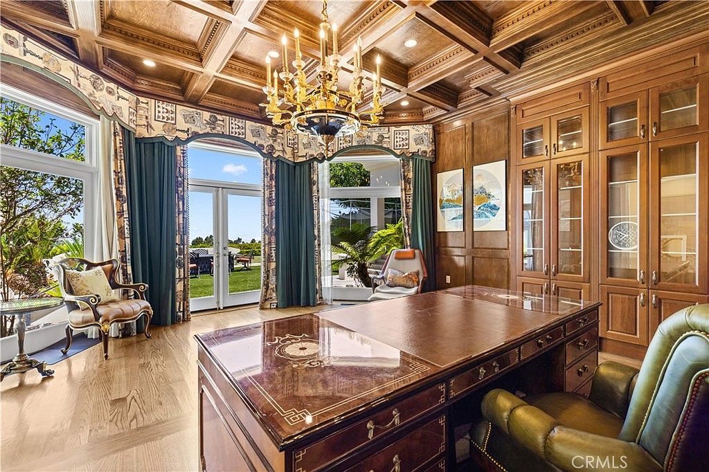 This elegant home office features rich wood-paneled walls, a coffered ceiling, and a large, polished desk as the central focal point. Large windows with blue drapery frame views of the outdoor landscape, while a classic chandelier adds a touch of sophistication to the room. The space is furnished with a leather office chair and an additional accent chair, creating a professional yet comfortable atmosphere.