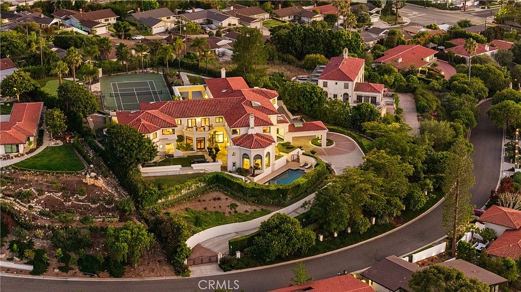 This high-angle aerial view captures a sprawling Mediterranean-style estate nestled within a lush, hilly residential neighborhood. The property features a prominent red-tiled roof, a private tennis court, and a swimming pool set against a backdrop of mature trees and manicured landscaping. A winding driveway leads to the residence, which is surrounded by terraced grounds and expansive outdoor living spaces.