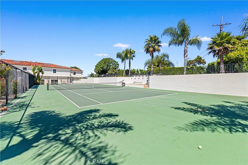 This image showcases a private, full-sized tennis court set within a spacious backyard, surrounded by a tall perimeter fence and a retaining wall. Several palm trees frame the court, adding a tropical aesthetic to the outdoor recreational space. The perspective is from the court level, looking toward the rear of the residence, which features a light-colored exterior and a tiled roof.