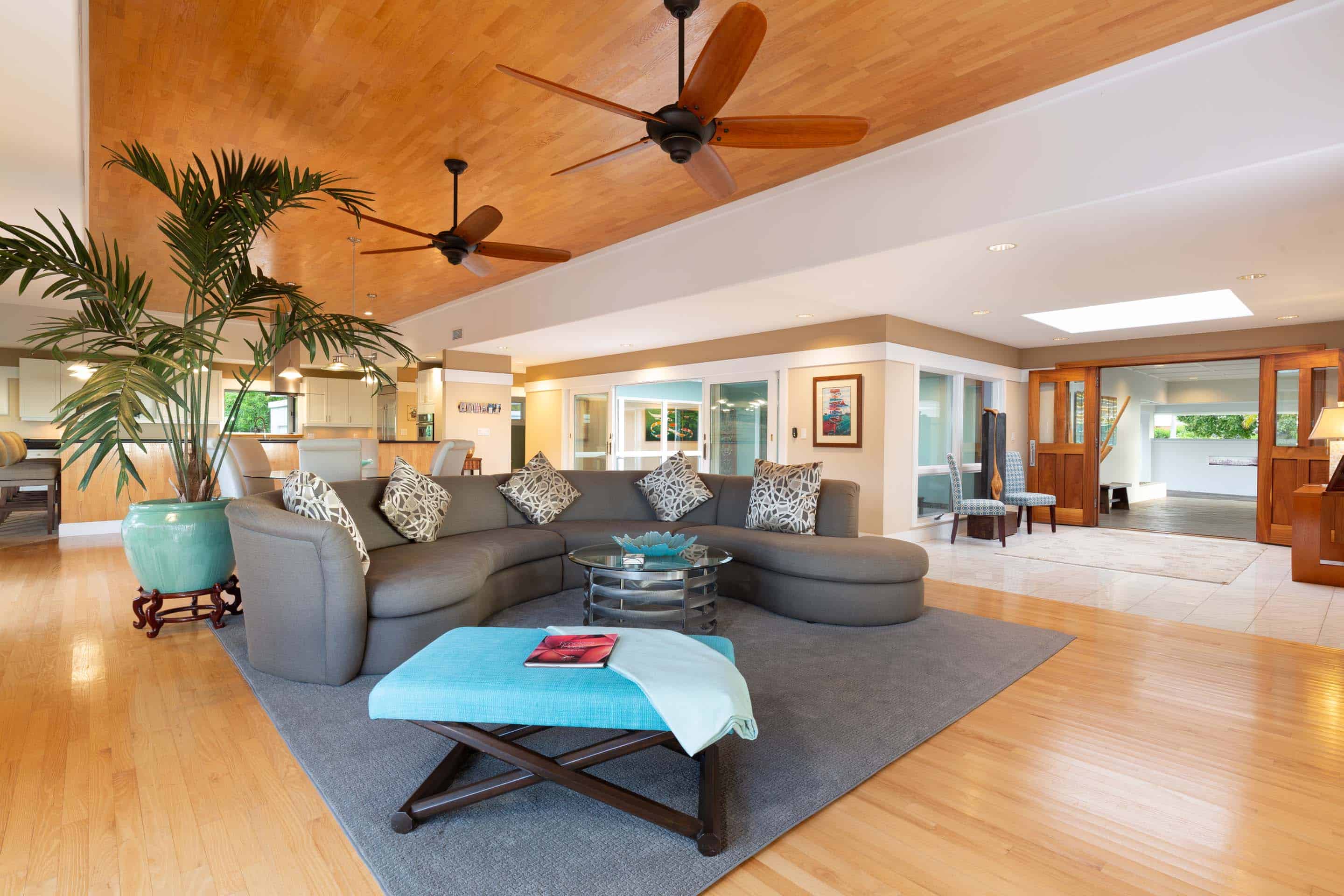 This is a spacious living room featuring a large, curved gray sofa with decorative pillows, a glass-topped coffee table, and a blue ottoman. The room is well-lit with natural light and features hardwood floors and a unique wood-paneled ceiling with ceiling fans. A large potted plant adds a touch of nature to the space.