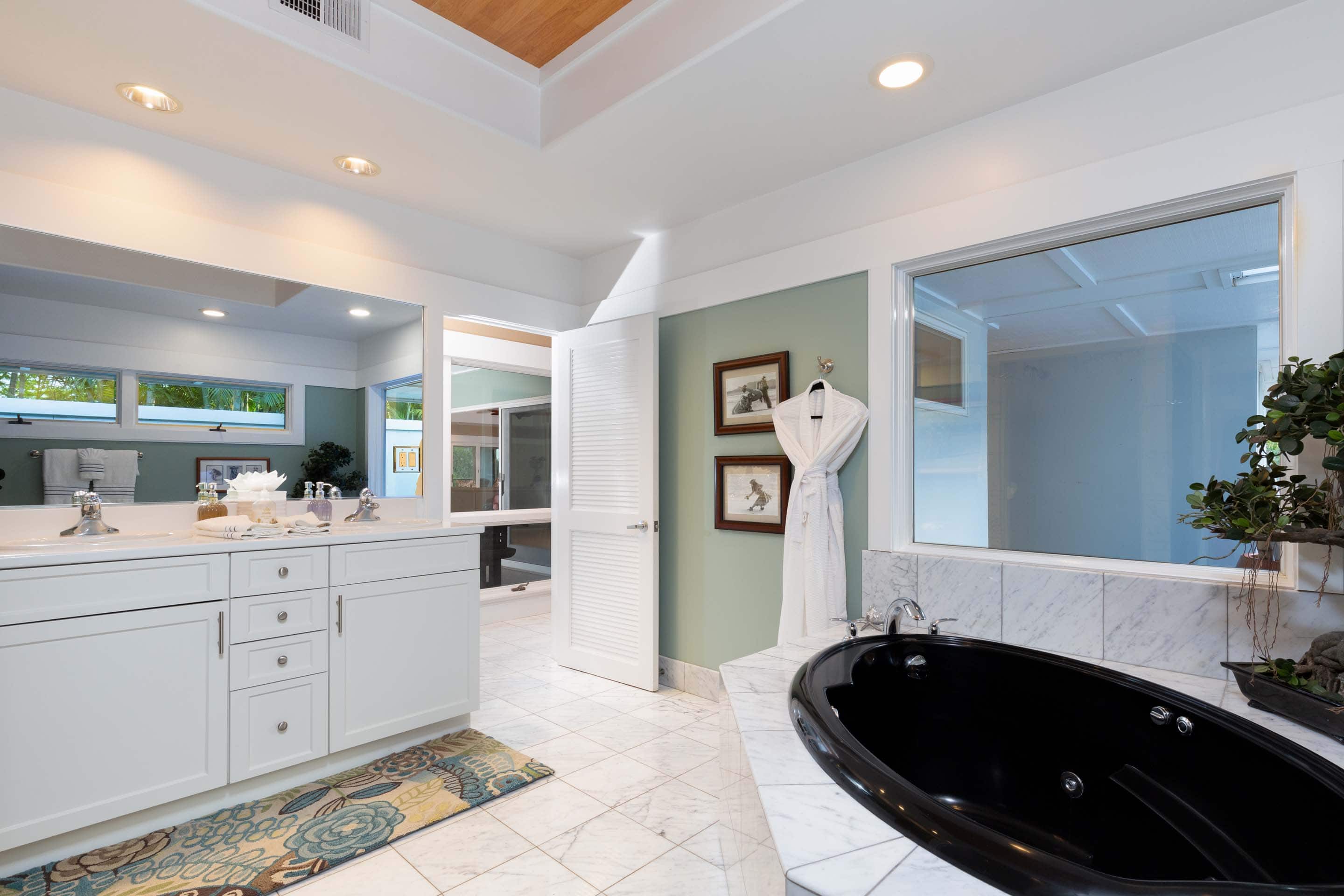 This is a bright and airy primary bathroom featuring a black soaking tub with marble surround, a white double vanity with ample storage, and a separate doorway leading to another room. The walls are painted a soft green, complemented by framed artwork and a white robe hanging on a hook. The overall impression is clean, luxurious, and well-maintained.
