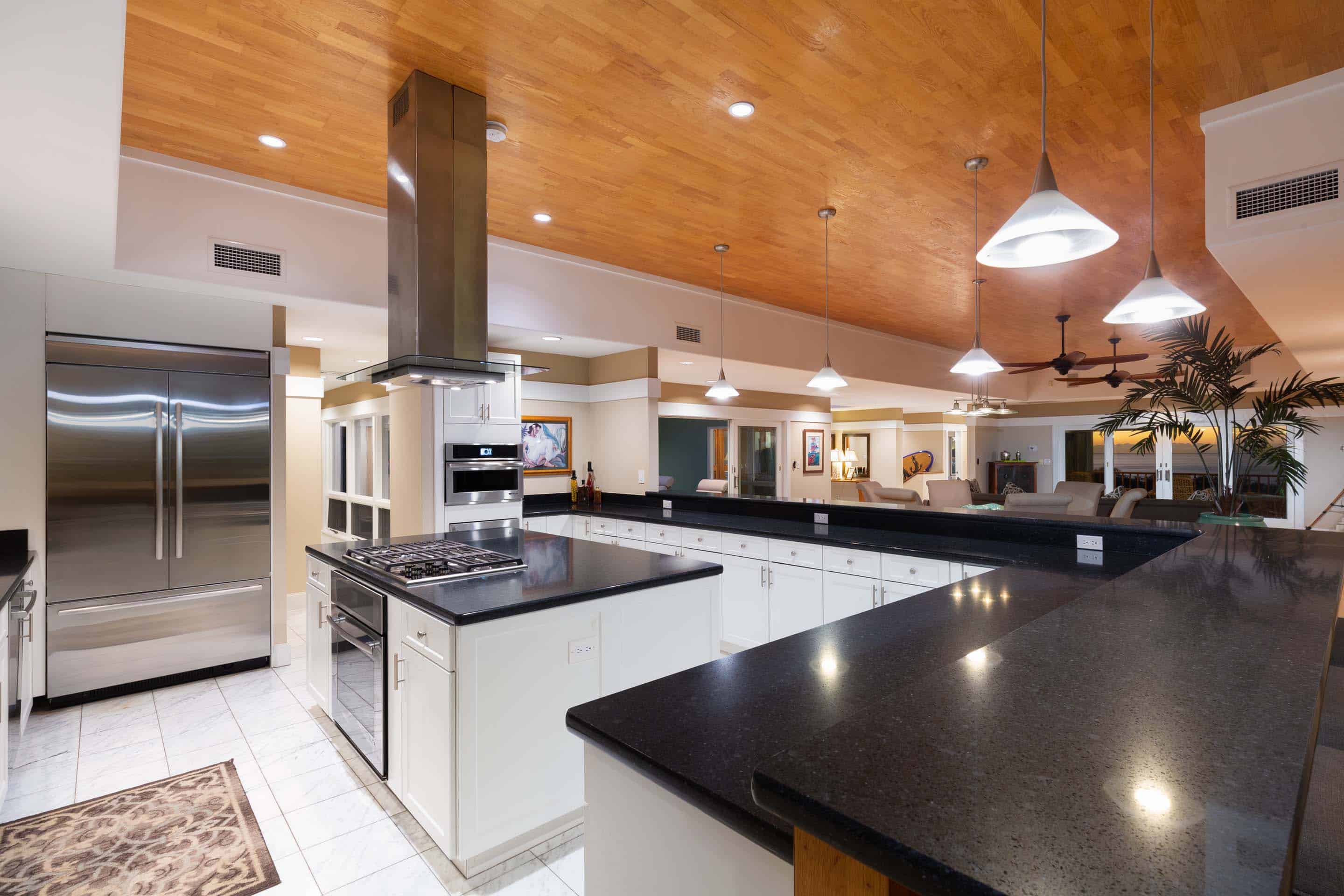 This is a well-lit kitchen featuring a large stainless steel refrigerator, white cabinetry with dark countertops, and a central island with a cooktop and oven. The kitchen opens to a living area, creating an open-concept space. The wooden ceiling adds warmth to the room, and pendant lights provide focused illumination over the island.