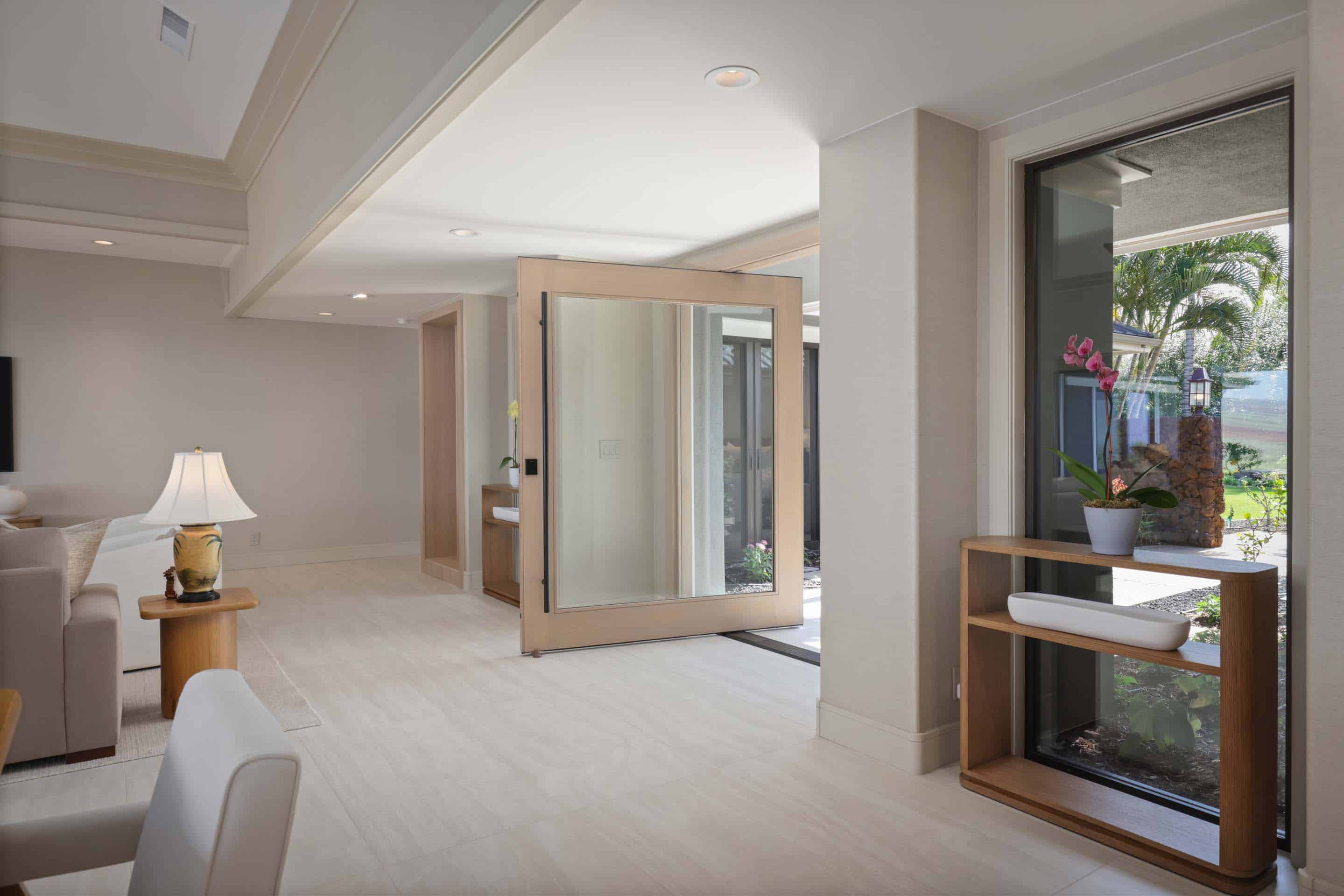 This interior shot showcases a bright and airy living room with a modern aesthetic. The space features light-colored tile flooring, neutral walls, and a large pivoting glass door that provides access to an outdoor area. A wooden console table with decorative items adds a touch of warmth, while the overall impression is clean, spacious, and inviting.