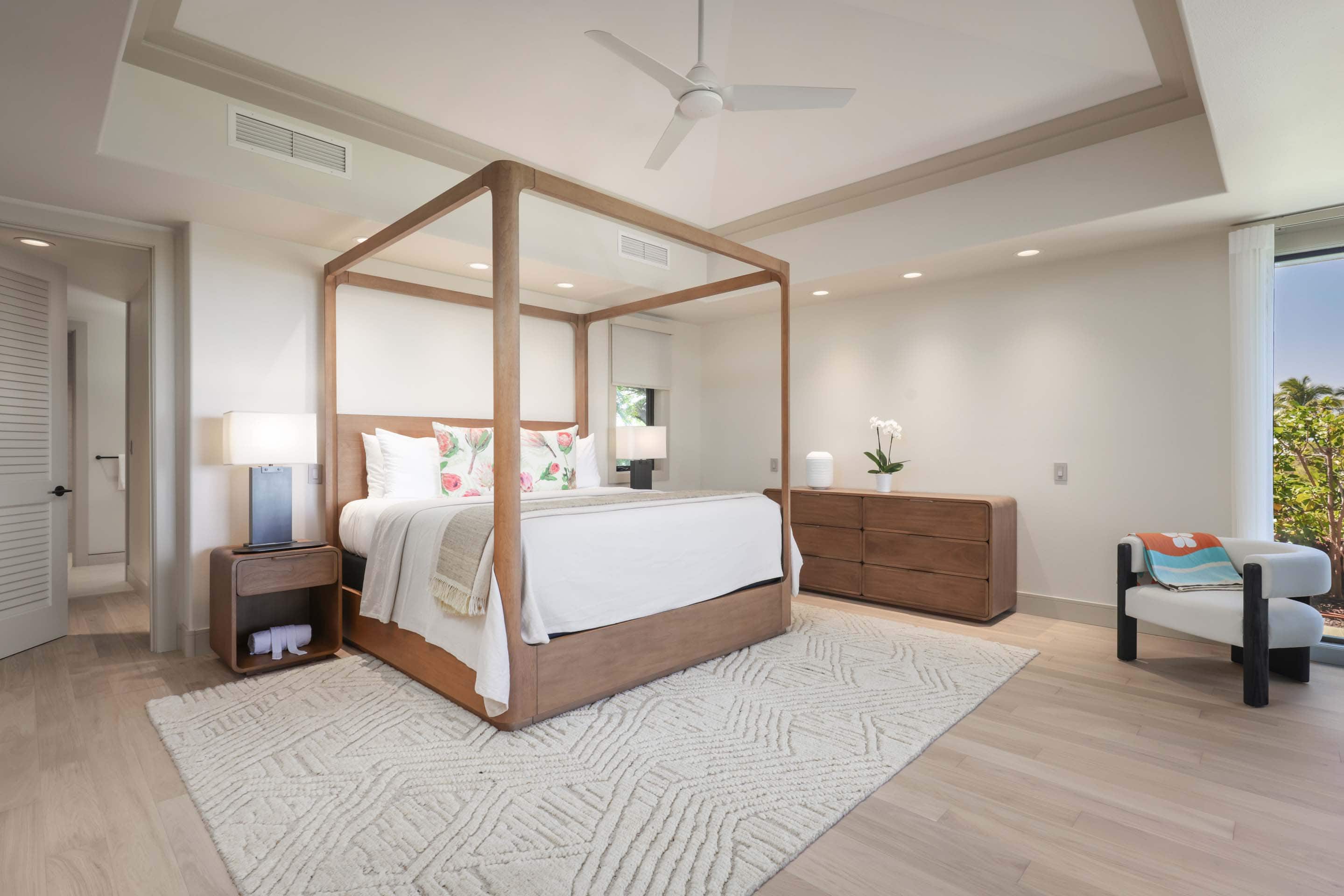 This is a bright and airy primary bedroom featuring a four-poster wooden bed with white linens and decorative pillows. The room is decorated in a minimalist style with light wood flooring, a neutral-colored rug, and a wooden dresser. A modern armchair sits near a large window, offering a view of the outdoors.
