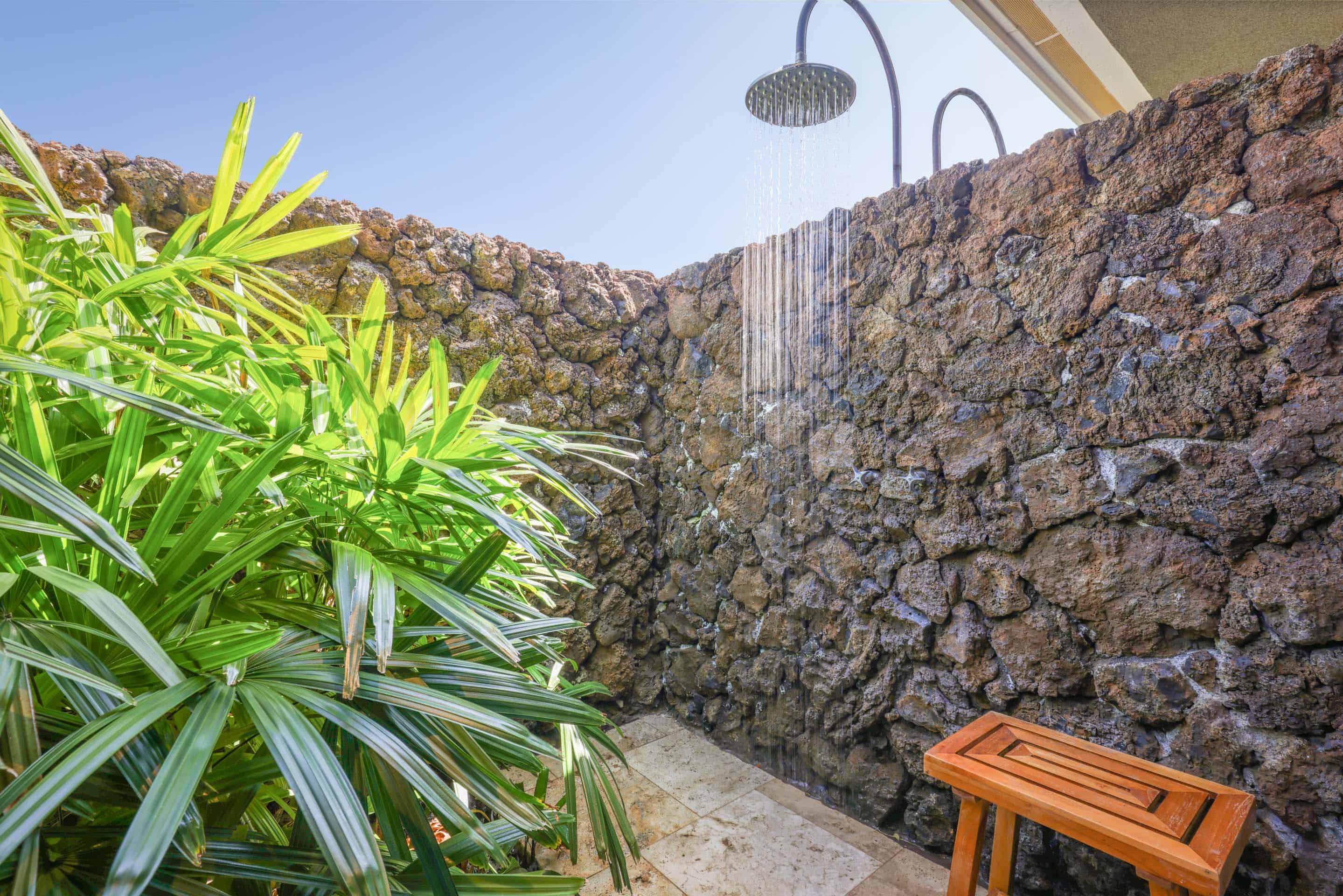 This image showcases an outdoor shower area, enclosed by a natural stone wall and lush tropical foliage. The showerhead is actively dispensing water, creating a refreshing ambiance. A small wooden bench sits nearby, adding a touch of rustic charm to the space.