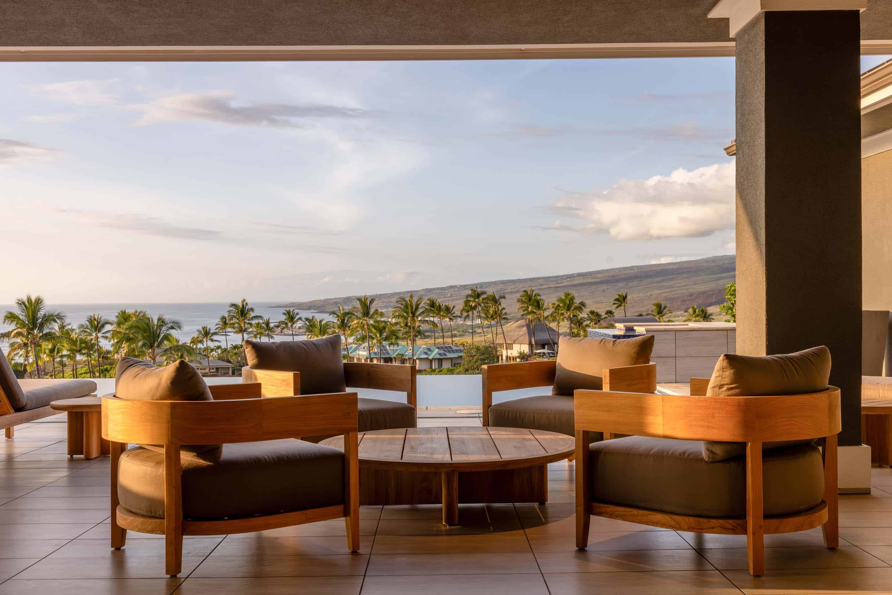 This image showcases a luxurious outdoor patio or balcony area with comfortable seating arranged around a wooden coffee table. The space offers stunning views of the ocean, palm trees, and distant coastline, creating a serene and inviting atmosphere. The design emphasizes relaxation and enjoyment of the natural surroundings, making it an ideal spot for entertaining or unwinding.
