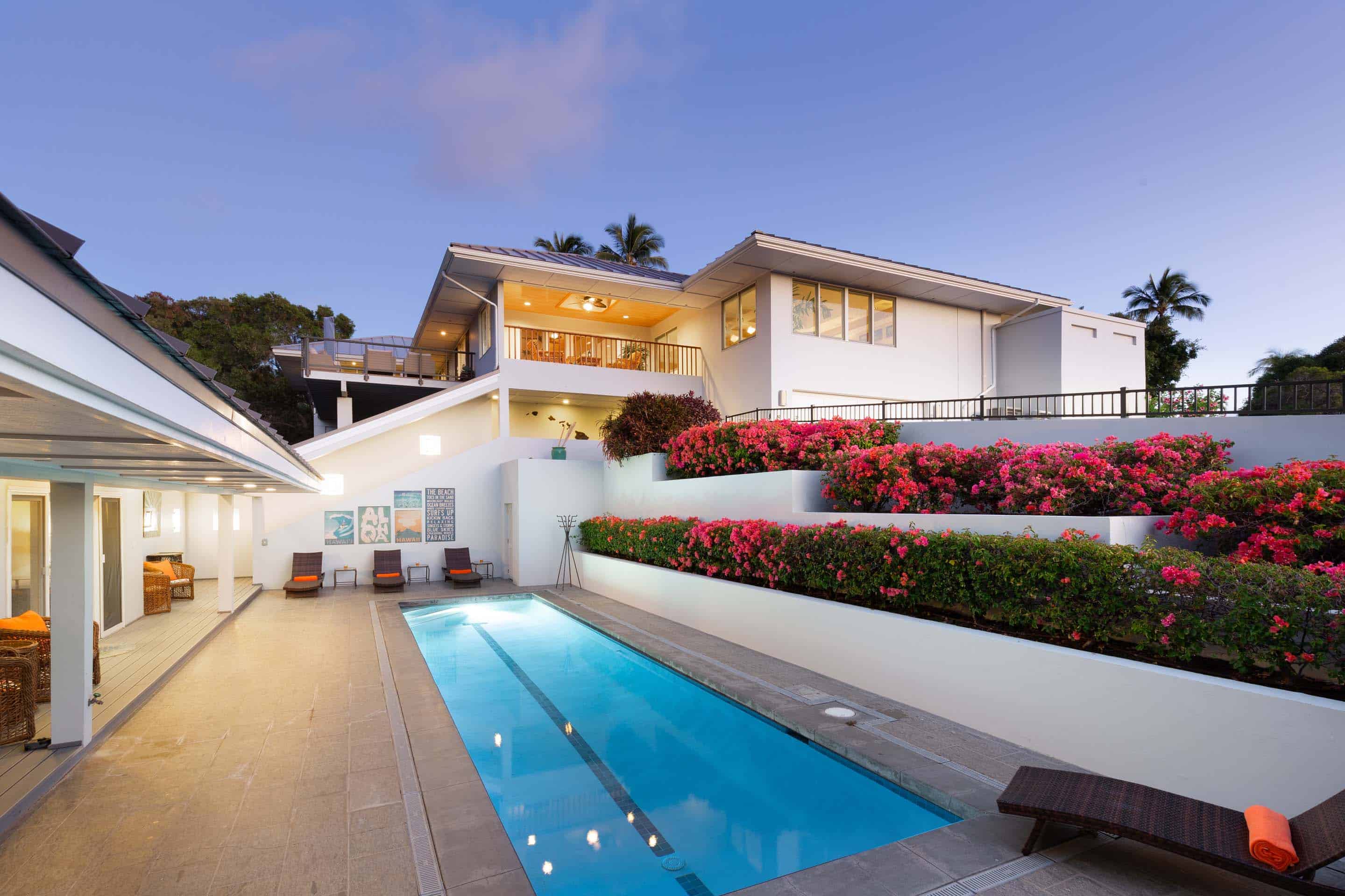 This image showcases a luxurious outdoor pool area of a modern home. The rectangular pool is surrounded by a tiled patio with lounge chairs, and a multi-tiered garden with vibrant pink flowers adds visual interest. The architecture of the house is contemporary, with clean white walls and multiple levels, creating an inviting and upscale atmosphere.