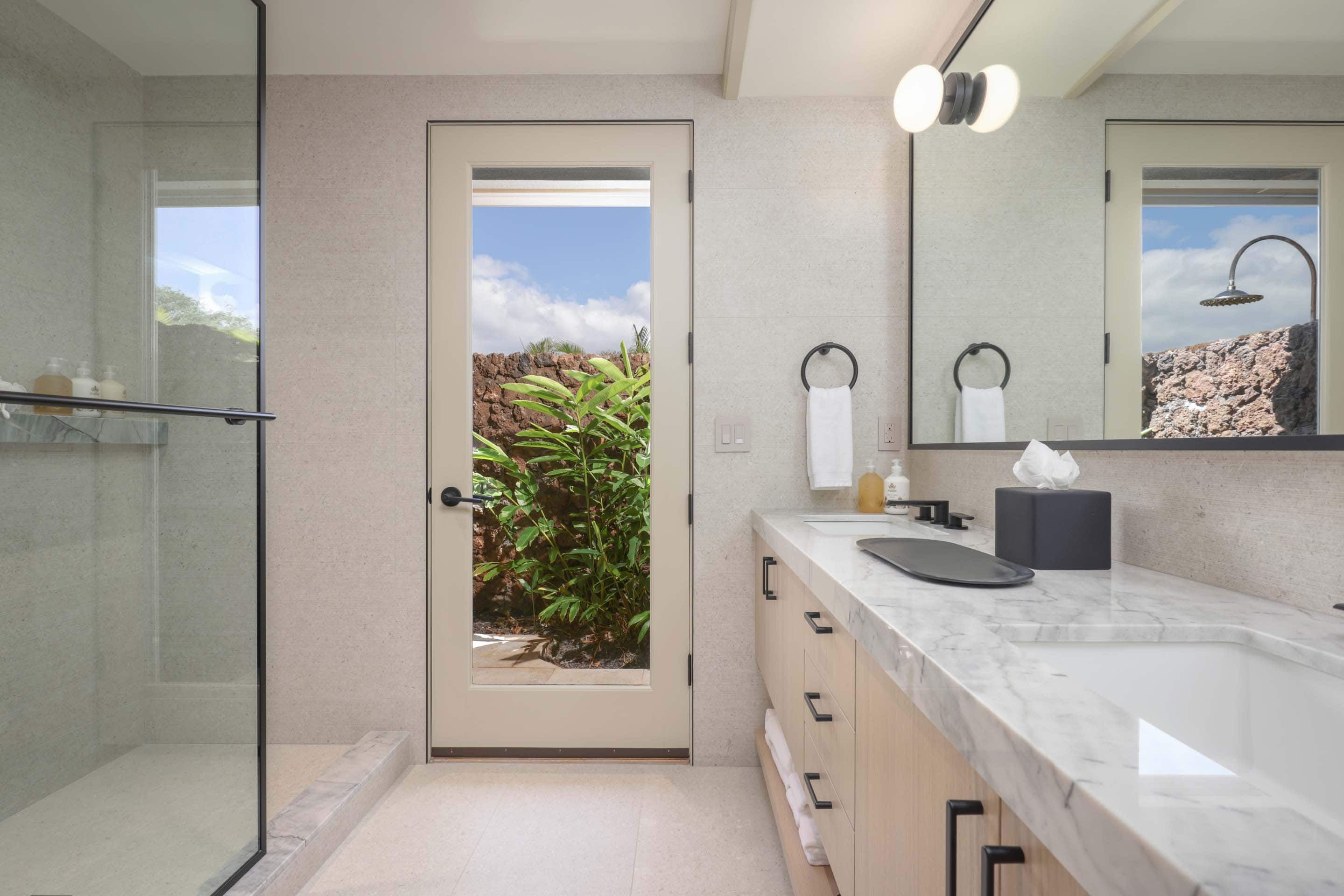 This is a bright and modern primary bathroom featuring a double vanity with a marble countertop and light wood cabinetry. A large mirror hangs above the vanity, reflecting natural light and a view of an outdoor garden through a nearby door. The bathroom also includes a glass-enclosed shower, creating a spa-like atmosphere.