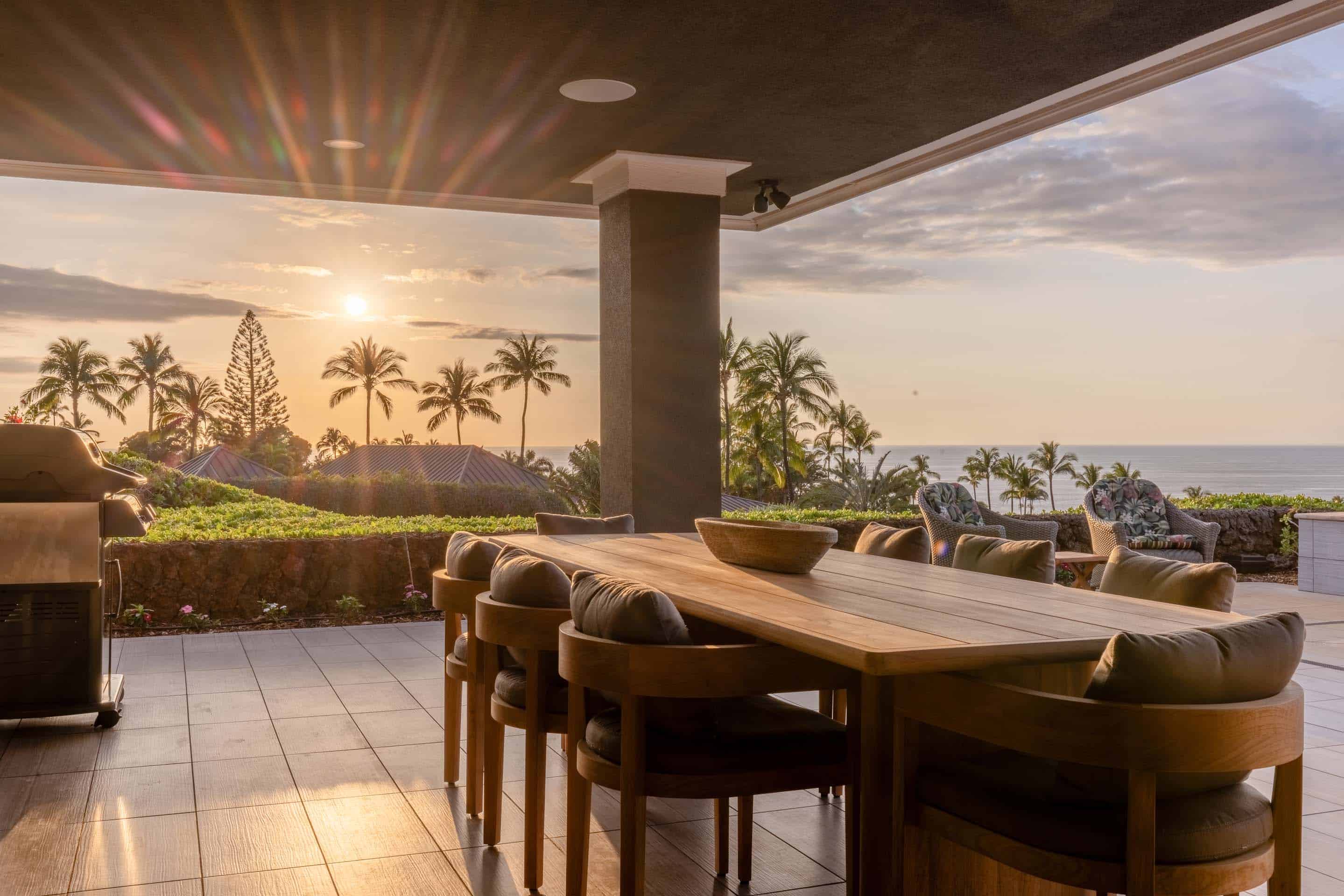 This image showcases a luxurious outdoor dining area on a patio or deck, featuring a large wooden table surrounded by cushioned chairs. The setting offers a stunning view of the ocean and palm trees at sunset, creating a serene and inviting atmosphere. A grill is visible on the left, suggesting an outdoor cooking space.