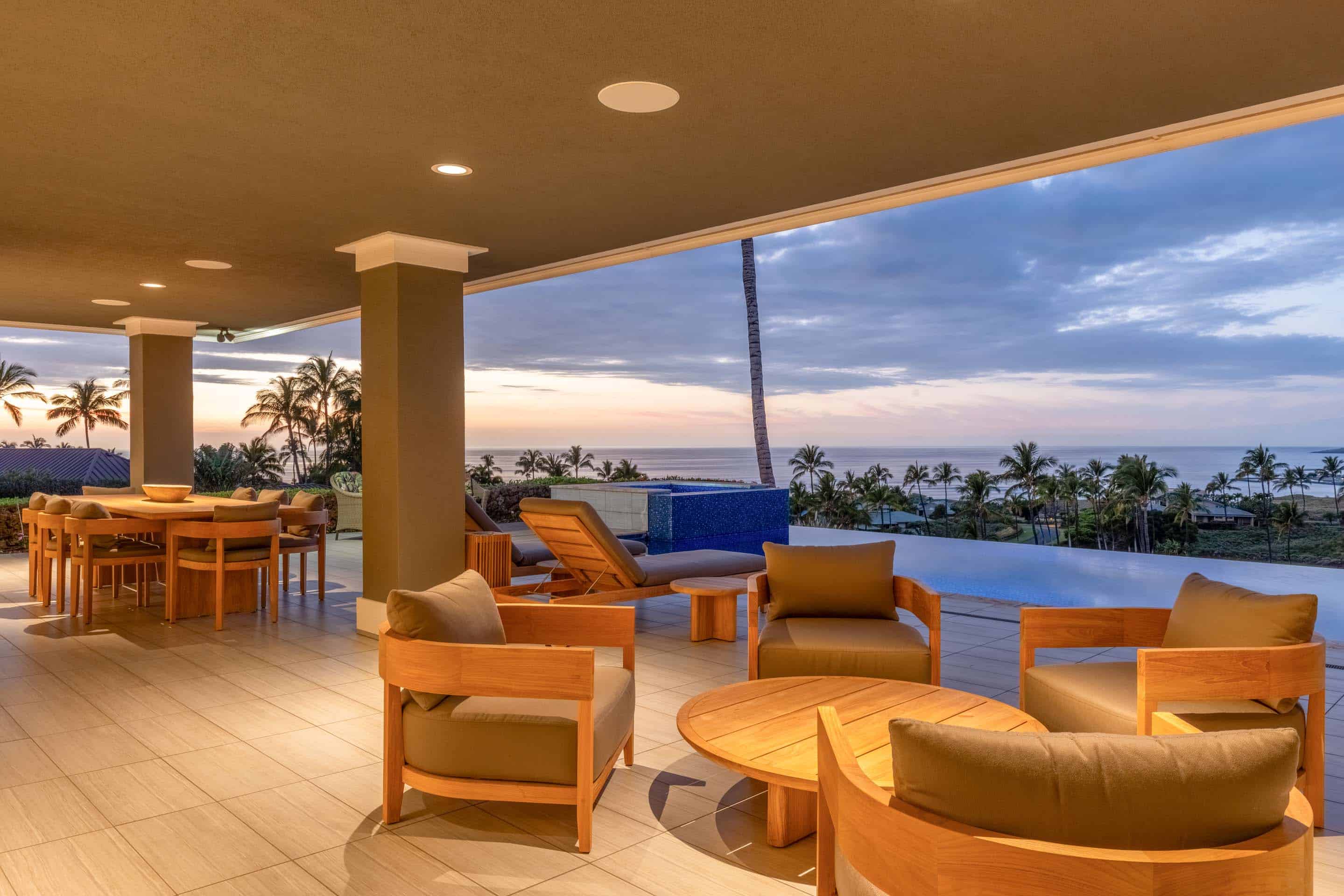 This image showcases a luxurious outdoor patio area with comfortable seating arrangements, including armchairs and a dining table, all crafted from wood. The patio overlooks a stunning infinity pool and the ocean, framed by palm trees and a vibrant sunset sky. The scene exudes relaxation and upscale living, perfect for enjoying the tropical scenery.