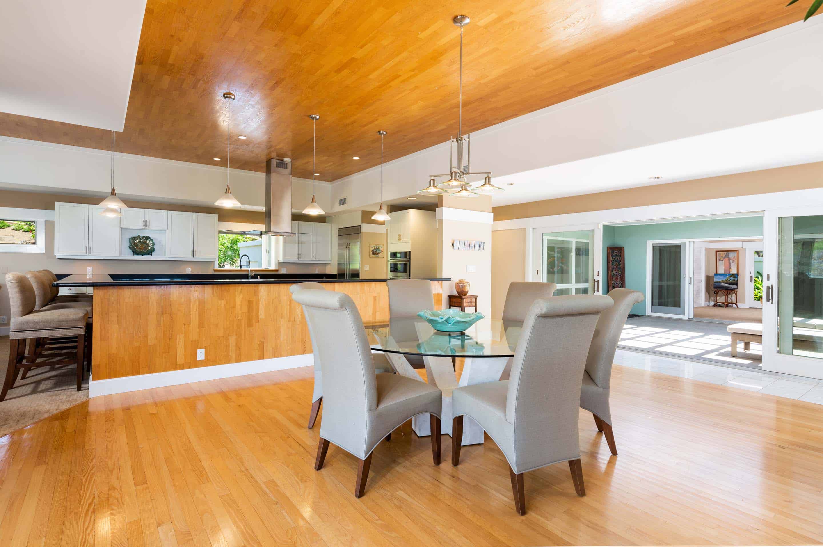 This is an interior shot of a dining room featuring a round glass table with six upholstered chairs. The room has hardwood floors and a unique wooden ceiling. Sliding glass doors lead to an outdoor area, enhancing the sense of space and light.