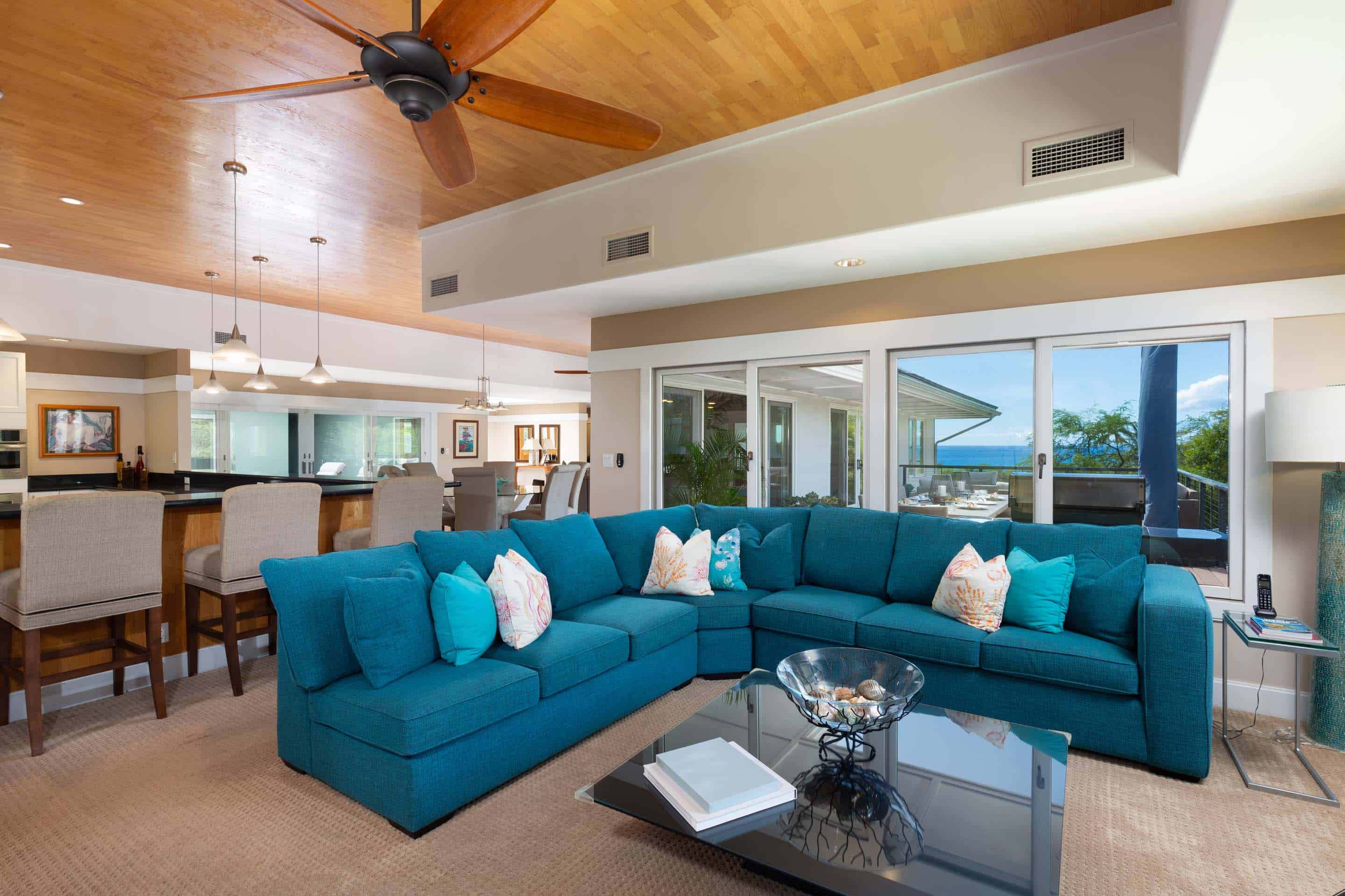 This is a spacious living room featuring a large, teal sectional sofa with decorative pillows, a glass-topped coffee table with decorative shells, and a view of the ocean through sliding glass doors. The room has a warm and inviting atmosphere, with a wood-paneled ceiling and neutral-toned walls. The perspective is from a medium shot, showcasing the entire living room.