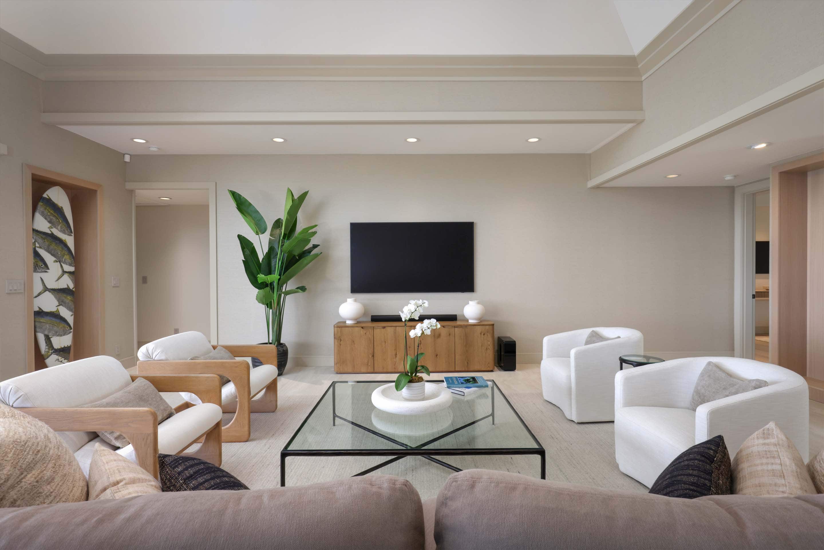 This is a bright and airy living room featuring a modern design with neutral tones. The room is furnished with comfortable seating, including armchairs and a sofa, arranged around a glass coffee table. A large television is mounted on the wall above a wooden cabinet, flanked by decorative vases, and a tall plant adds a touch of nature to the space.