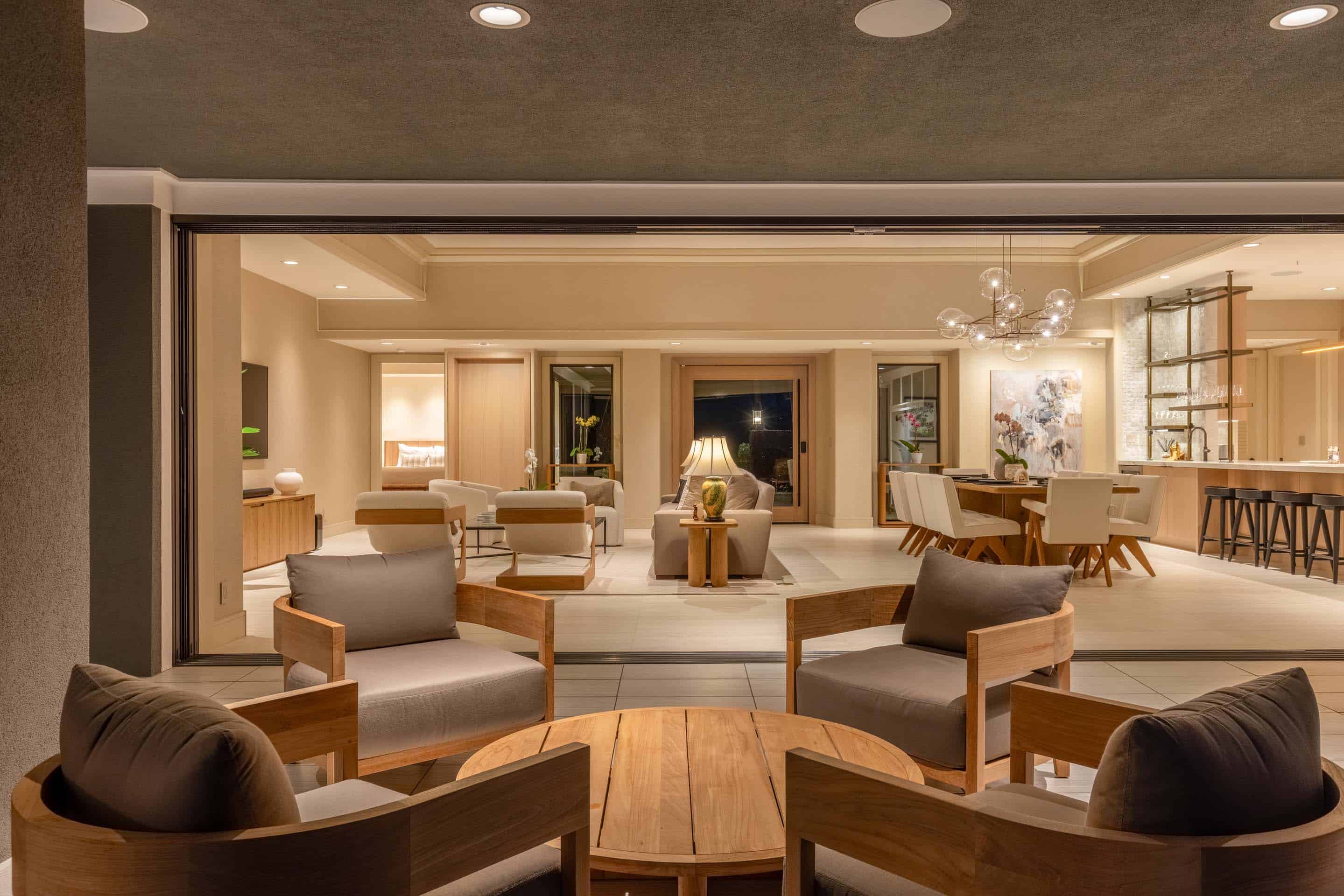 This interior shot showcases a luxurious living space that seamlessly blends indoor and outdoor living. The open floor plan features comfortable seating arrangements with wooden-framed chairs and a round coffee table, leading into a dining area and kitchen with bar seating. Large sliding glass doors provide ample natural light and access to an outdoor area, enhancing the sense of spaciousness and modern elegance.