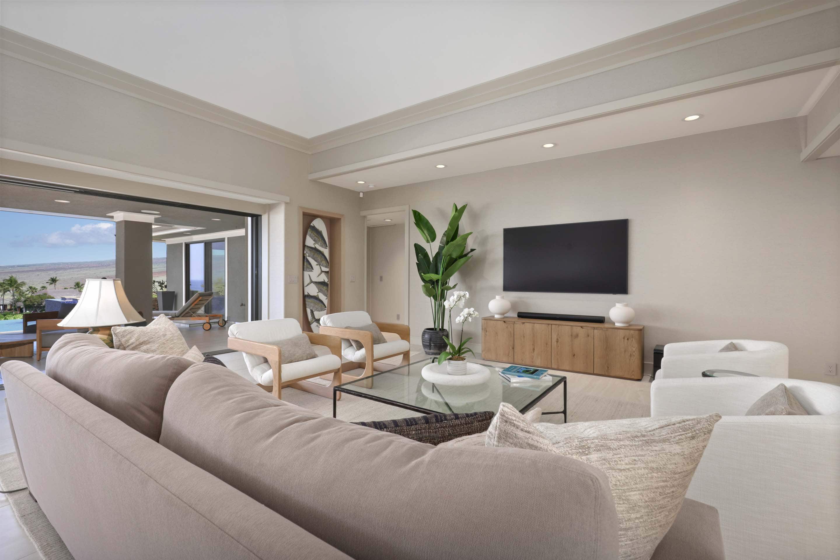 This is an interior shot of a living room featuring a large beige sofa, two white armchairs, and a glass coffee table. A large television is mounted on the wall above a wooden console, and a sliding glass door leads to an outdoor patio area. The room has a modern, minimalist style with neutral colors and natural light.