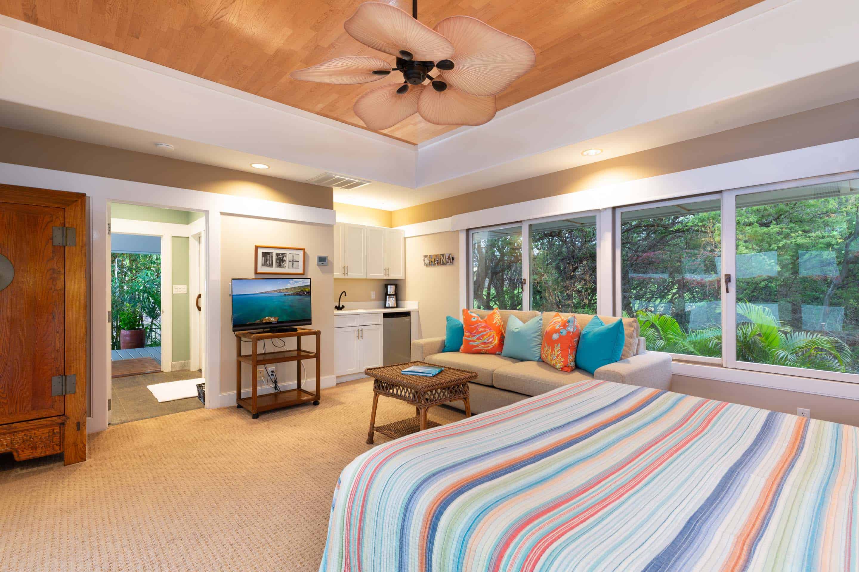 This is a cozy bedroom featuring a bed with striped bedding, a comfortable sofa with decorative pillows, and a wicker coffee table. The room has a kitchenette area with white cabinets and a small refrigerator. Large windows offer a view of lush greenery, creating a relaxing and inviting atmosphere.