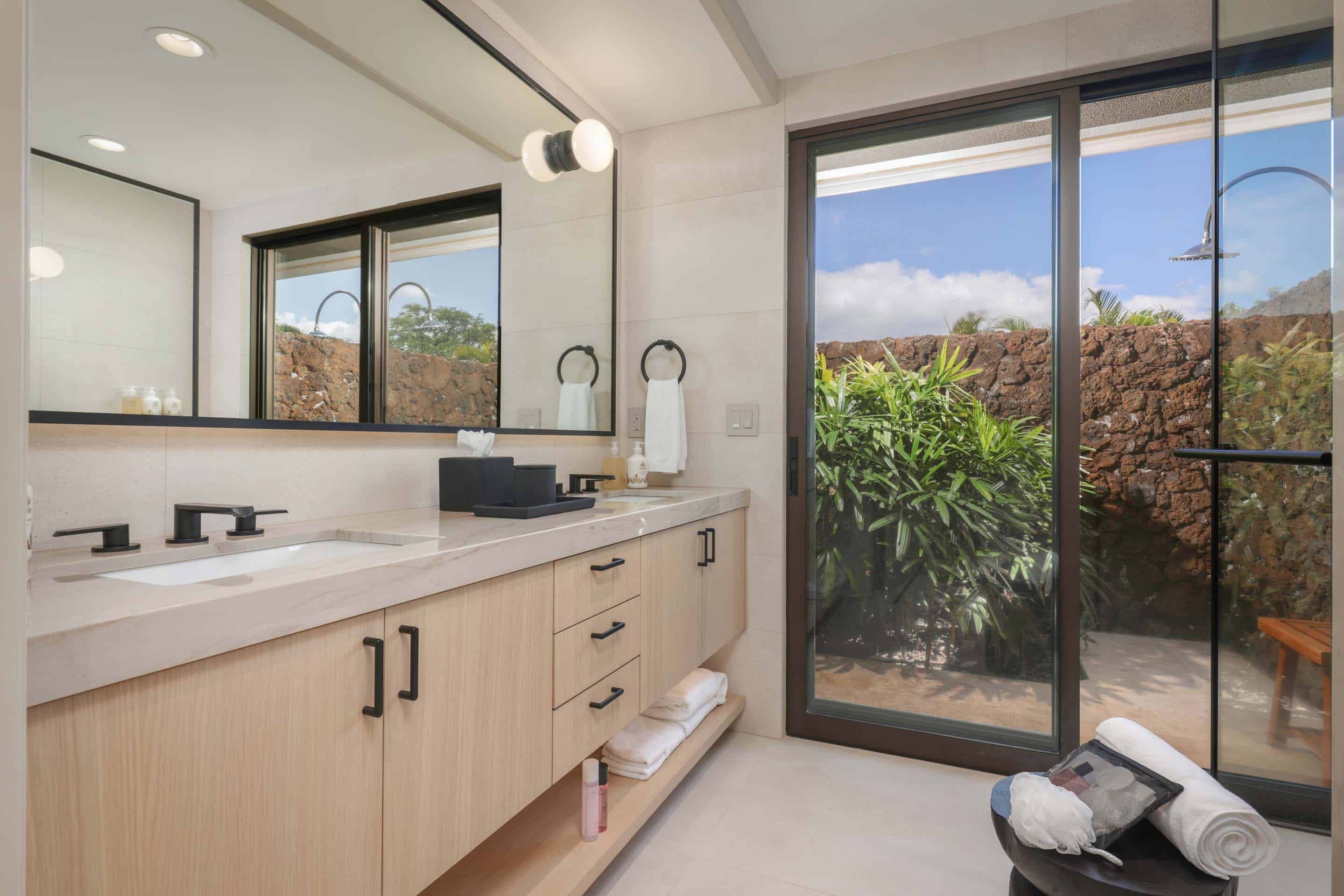 This is a bright and modern primary bathroom featuring a double vanity with light wood cabinetry and black hardware. A large mirror reflects the natural light, and a sliding glass door leads to an outdoor shower area surrounded by lush greenery and a stone wall. The overall impression is clean, luxurious, and spa-like.