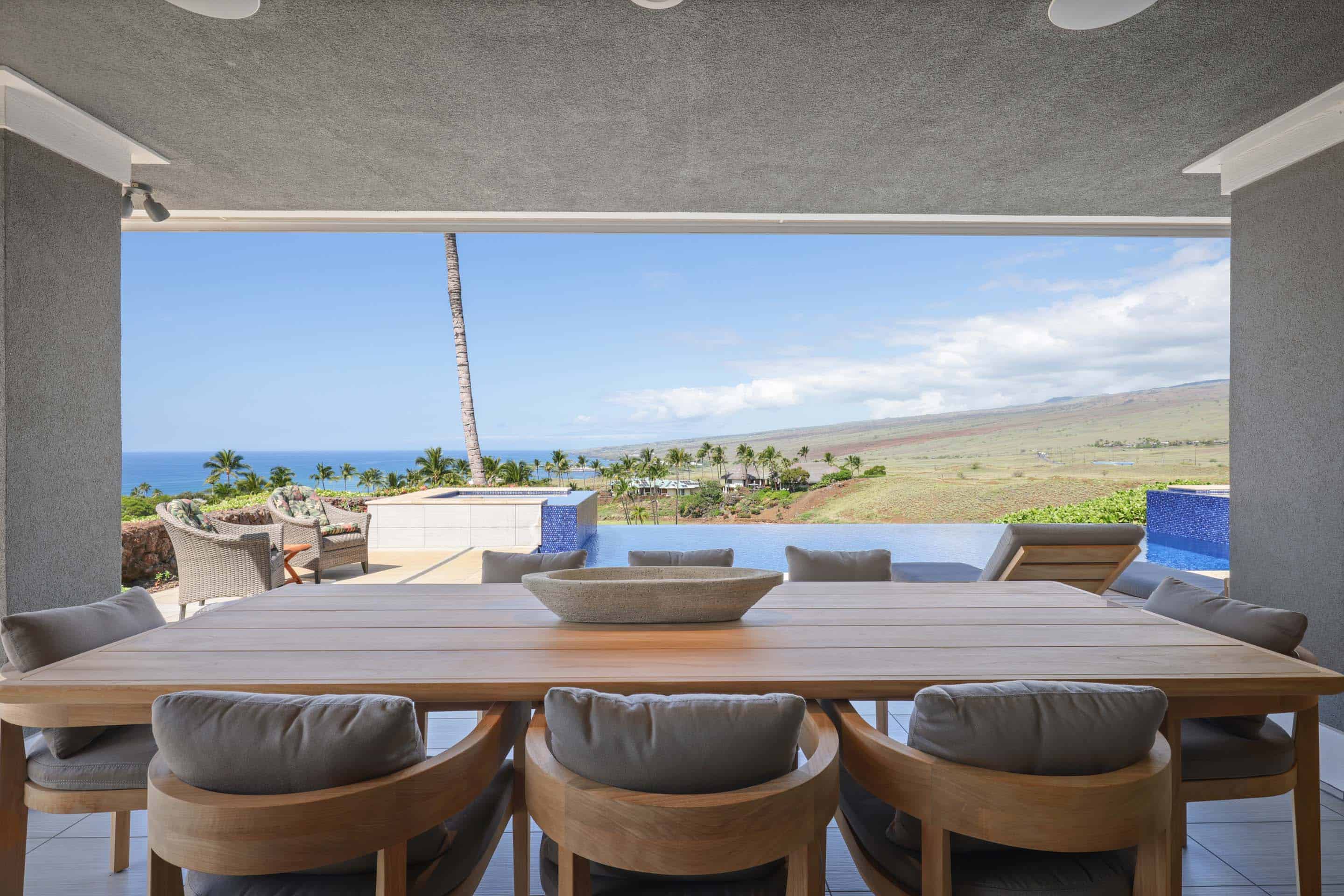 This image showcases a luxurious outdoor dining area on a patio or balcony, featuring a large wooden table surrounded by cushioned chairs. The space overlooks an infinity pool and offers stunning views of the ocean and landscape, creating a serene and upscale ambiance. The perspective is from inside the patio looking out.