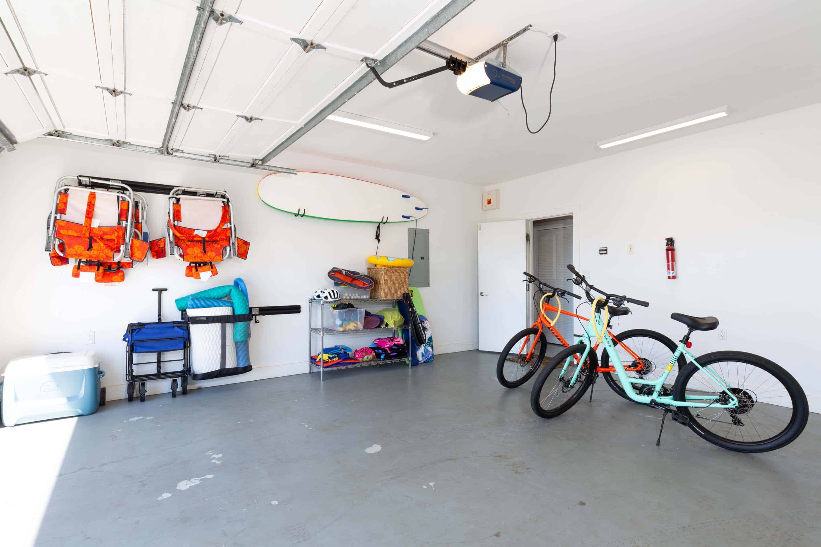 This is a well-organized garage featuring a variety of storage solutions. Beach chairs and a surfboard are mounted on the wall, while a metal shelving unit holds various items. Two bicycles are parked in the center of the garage, and the floor is a clean, neutral gray. The perspective is from the center of the garage looking towards the back wall.