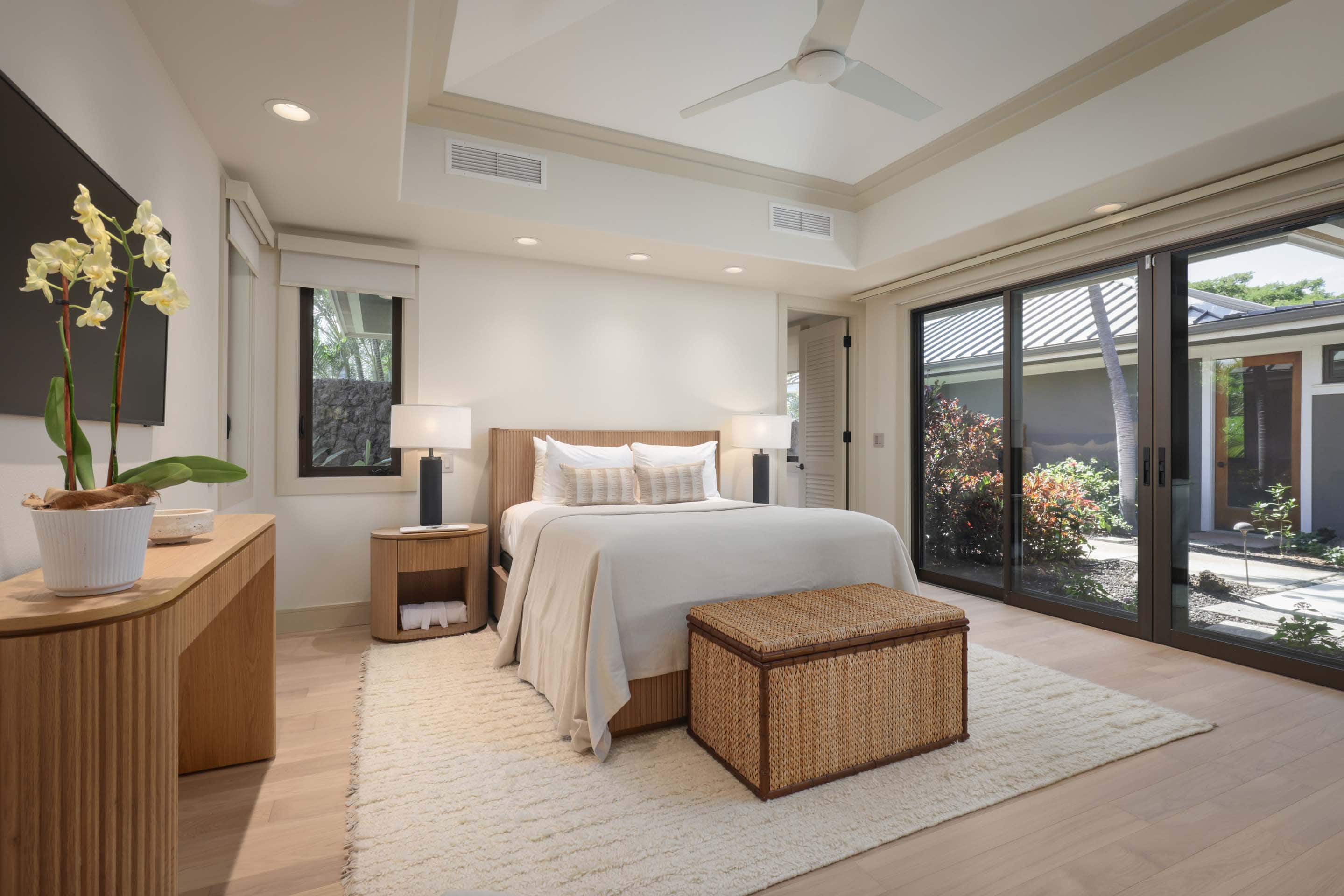 This is a bright and airy primary bedroom featuring a neutral color palette. The room includes a wooden bed frame with light-colored bedding, a woven storage bench at the foot of the bed, and matching wooden nightstands with lamps. Large sliding glass doors provide access to an outdoor area, enhancing the room's connection to nature.