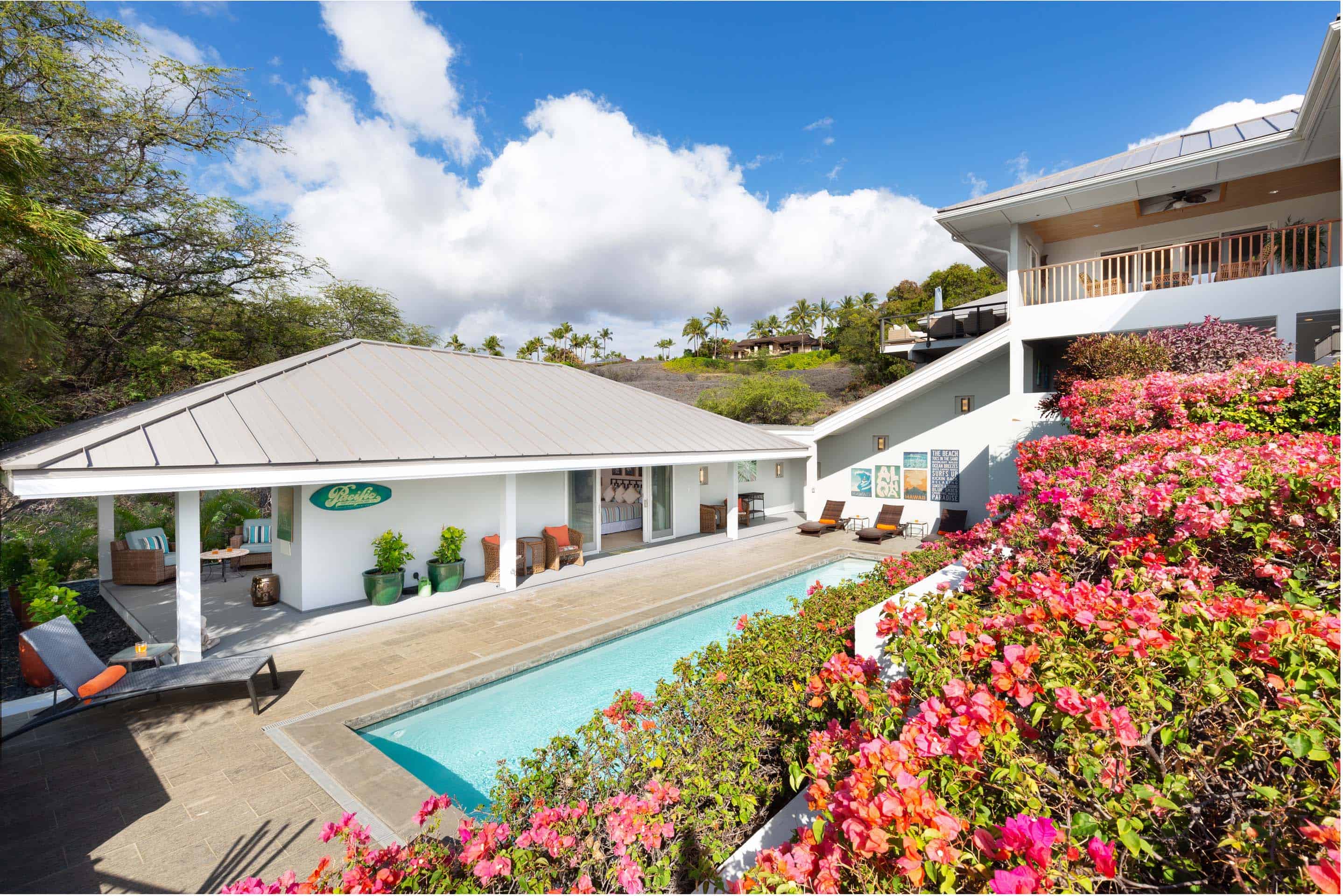 This image showcases a luxurious outdoor pool area, framed by vibrant pink bougainvillea. The pool is surrounded by a paved patio with lounge chairs, and a covered outdoor living space is visible in the background. The architecture is modern and clean, with a second story balcony visible on the right.